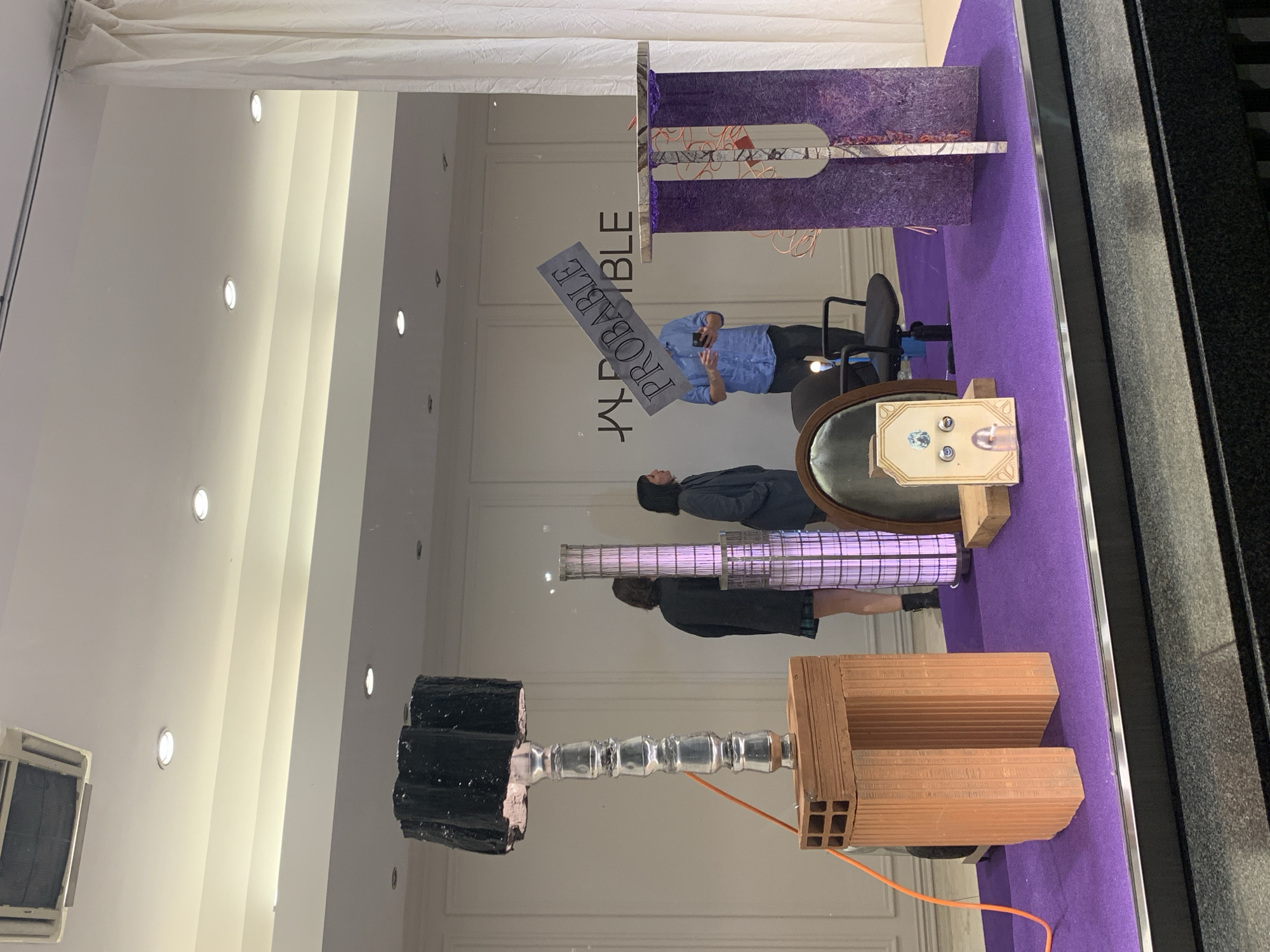 A retail window display exhibits an assortment of objects against a purple backdrop, likely within a building in Buenos Aires, Argentina, judging by architectural cues and cultural elements not apparent in this frame. The display incorporates a black cylindrical object affixed to a metallic, ribbed structure, alongside a terracotta brick assembly. Nearby, a cylindrical, cage-like structure stands next to a framed circular mirror with objects resting on a small wooden pedestal. Reflections of two people are visible in the glass, alongside a man holding a sign with the text "PROBABLE". The architectural details of the store's interior, like ceiling lights and paneling, are also reflected in the glass.