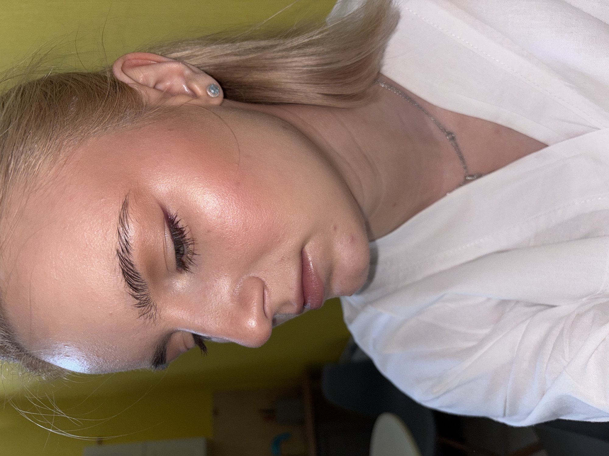 A close-up, profile view of a young Caucasian woman with blonde hair and fair skin. She is wearing a white shirt and a silver necklace with a small pendant. She has on makeup, including eyeliner and blush. A single stud earring with a light blue stone is visible on her right earlobe. The background is a blurred yellow wall. The image appears to be taken indoors, possibly in an apartment or studio. There is no discernible location in Saint Petersburg, Russia, or any activity taking place.