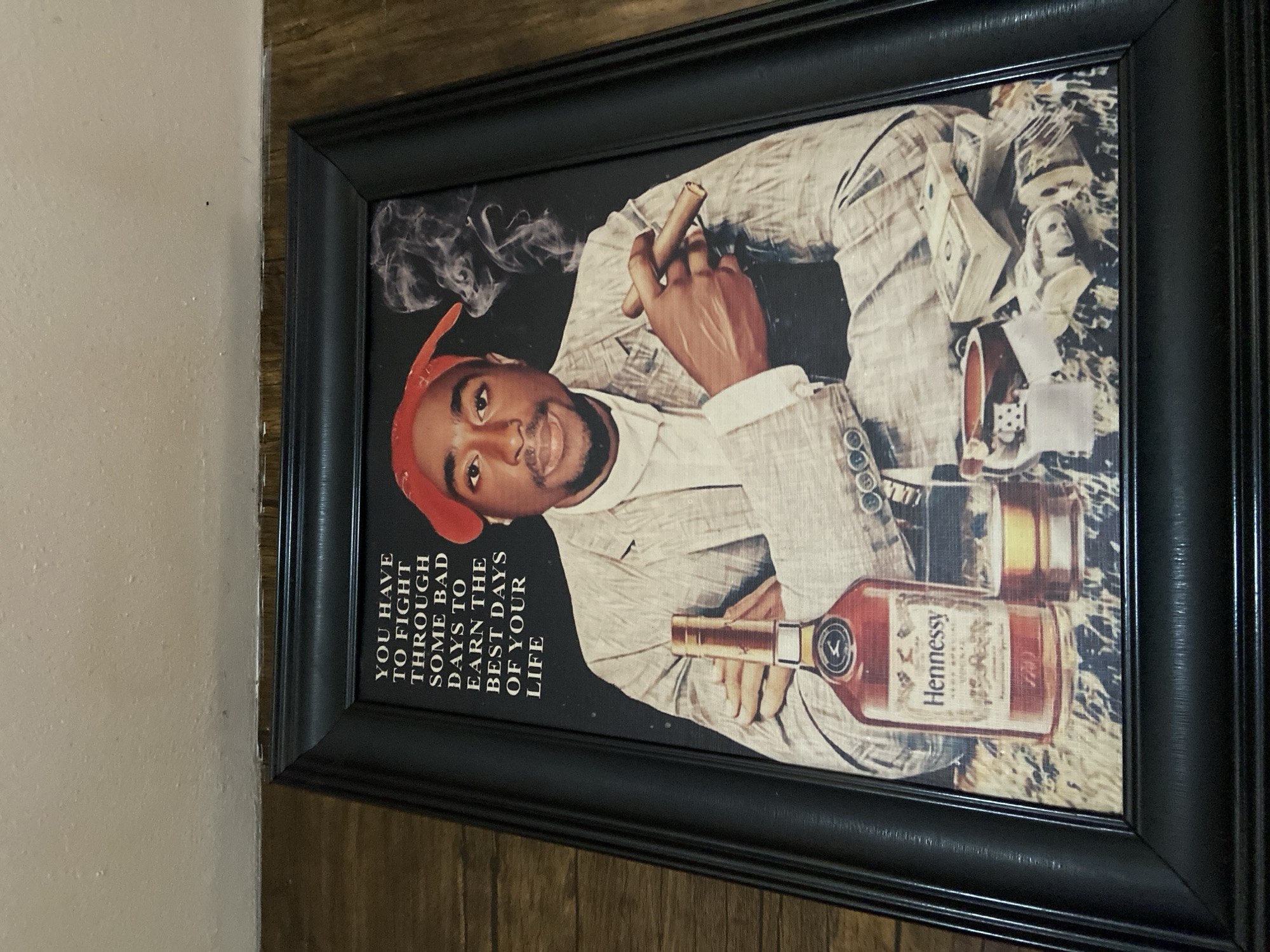 A framed picture, mounted on a wall with a textured, light-colored surface and a darker wooden-like surface, is the central subject of this image. The picture features a prominent depiction of a man, resembling the late rapper Tupac Shakur, wearing a red bandana, a white shirt, and a light-colored suit jacket. He is looking directly at the viewer with a slight smile.
In his right hand, the man holds a lit cigar, from which smoke visibly curls upwards. To his right, stacks of what appear to be U.S. $100 bills are arranged, suggesting wealth or success. Below the cash, a glass and a distinctive bottle of Hennessy cognac are displayed. The background within the picture is dark and somewhat abstract, emphasizing the central figure and accompanying elements. The entire artwork is enclosed in a thick, black frame.
On the left side of the artwork, a motivational quote is printed vertically in white text, stating: "YOU HAVE TO FIGHT THROUGH SOME BAD DAYS TO EARN THE BEST DAYS OF YOUR LIFE". The label on the cognac bottle clearly displays the brand name "Hennessy" along with "V.S.O.P" and "Cognac". The image captures the framed picture at a slight angle, indicating it was taken from an overhead perspective. The details of the wall and the frame's texture are also visible, hinting at the physical setting where the picture is displayed.