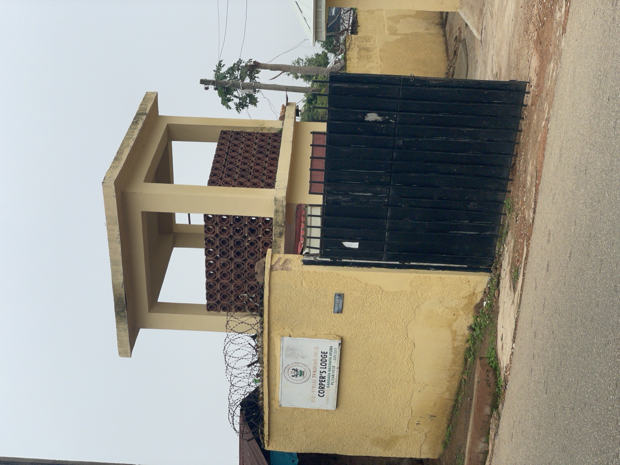 The image depicts the entrance to "Corper's Lodge" in Jos, Nigeria. The lodge is a building with a yellow, stuccoed exterior. A prominent feature is a two-tiered structure with a concrete roof and yellow pillars, topped with a decorative screen of circular terracotta elements. Barbed wire is installed on top of a section of the wall. A large black metal gate stands at the entrance, partially ajar. A sign on the yellow wall reads "INDUSTRIAL TRAINING FUND CORPER'S LODGE COURTESY GANDADA NACHAIYA AYUBA PL/16B/4578 2016-2017." A smaller sign on the gate also reads " CORPER'S LODGE". A paved road is visible in the foreground. A utility pole with electrical wires is situated to the right of the building, with a tree next to it. The sky is overcast. The overall impression is that of a secure compound, likely housing youth corps members.