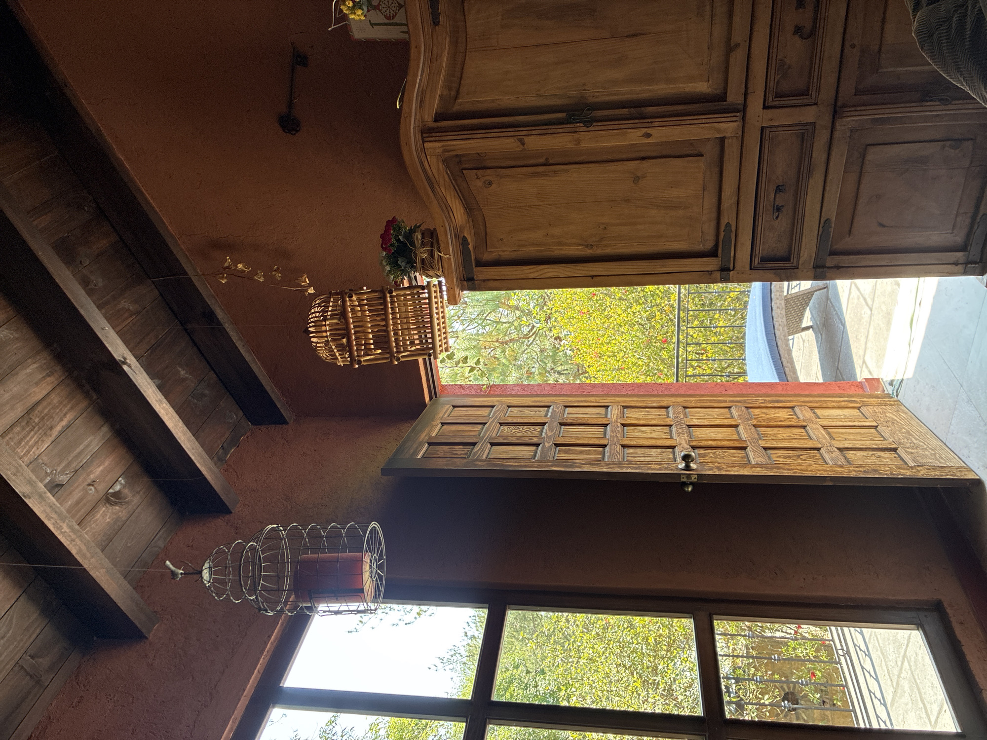 The scene is an interior view of a building, seemingly in Cañada de Madero, Mexico, as the prompt suggests. The perspective is unusual, looking upwards at an angle. The ceiling is a mix of reddish-brown textured surfaces and exposed dark wooden beams. Two hanging ornaments are visible: a wicker basket with flowers, and a metal cage-like structure containing a red cylindrical object. The lighting appears to be coming from both an open door leading to an outdoor patio area with green foliage and from windows showing a green landscape. A large wooden cabinet or cupboard dominates the upper portion of the image. A dark wooden door with a grid pattern is visible near the window, partially open.