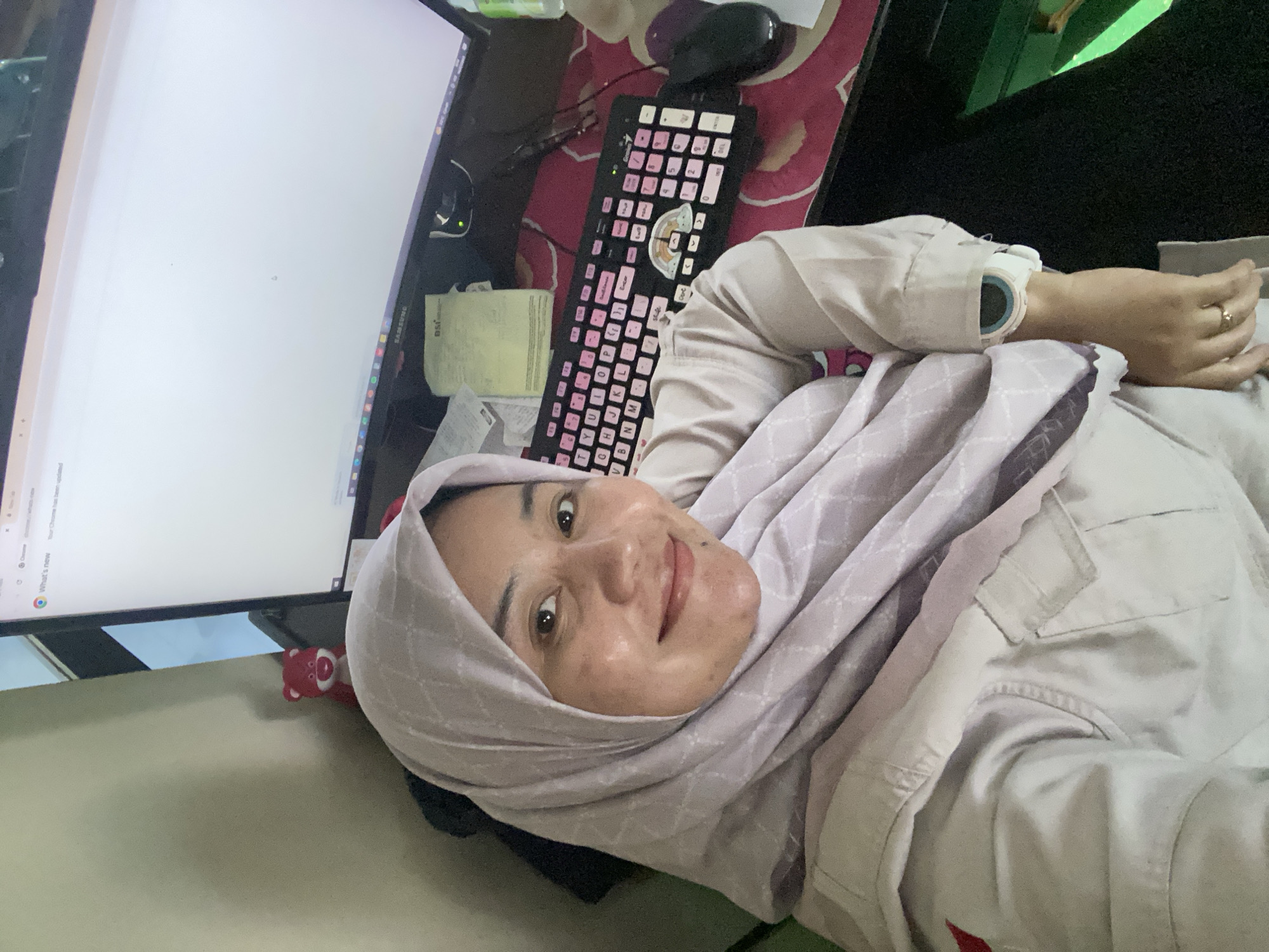 A woman wearing a light purple hijab and a light-colored uniform is seated at a desk in what appears to be an office or study in Banjarbaru, Indonesia. She is taking a selfie with her phone, as indicated by the angle of the shot. A computer monitor, keyboard with pink keycaps, and a mouse are visible on the desk. A small red bear figurine is perched on the edge of the monitor. The woman has a subtle smile and her right hand rests on the desk, adorned with a ring. Her left arm, wearing a white smartwatch, is also visible. The background shows a light-colored wall and a portion of a window with a dark frame. The overall scene suggests a casual moment during a workday.