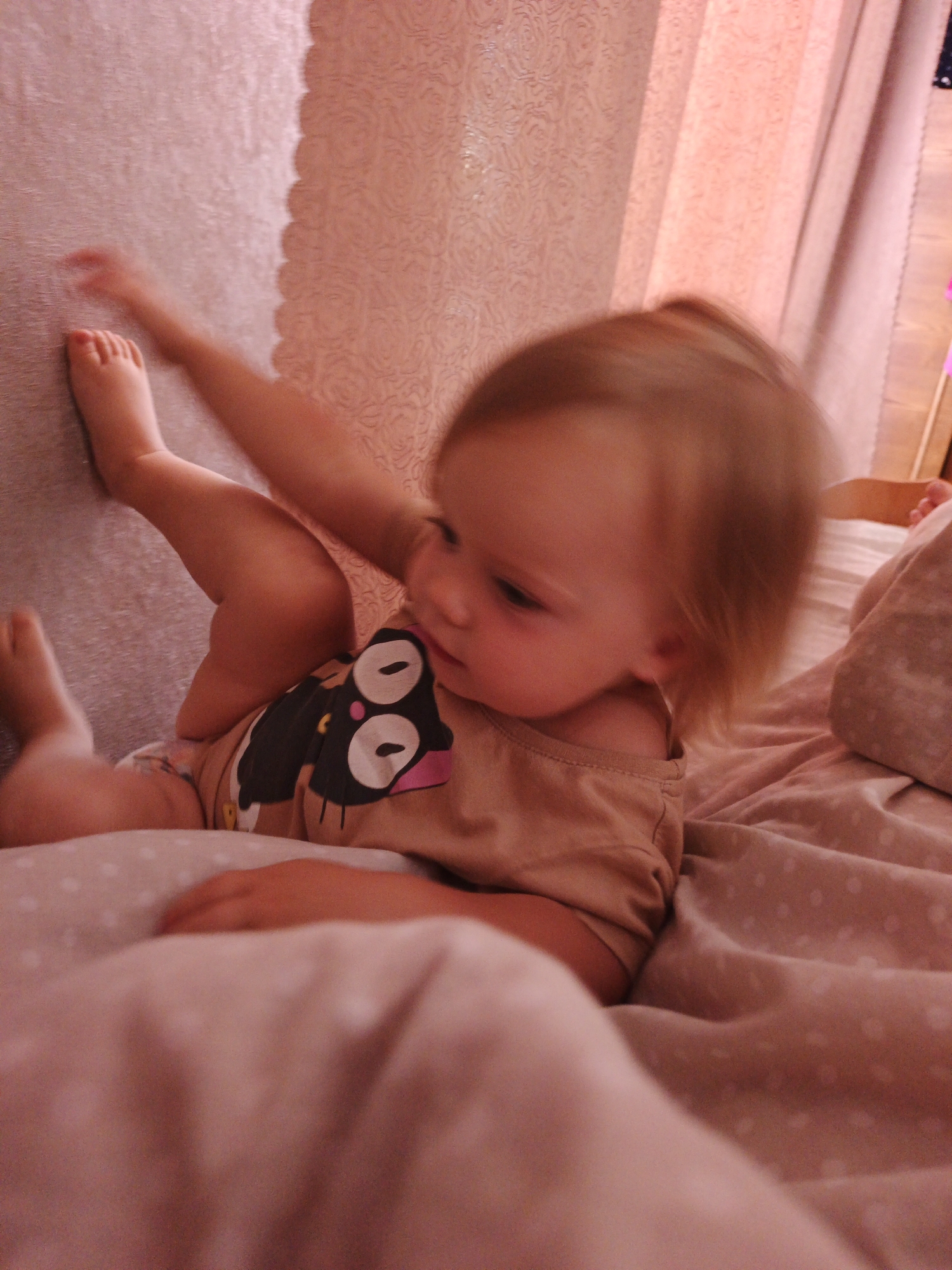 The image depicts a young child lying on a bed with pink polka-dot bedding. The child is wearing a tan onesie with a graphic of a black cat face. The child's left leg is extended upwards, and their right leg is bent. The child's head is turned to the side, with their hair blurred due to motion. A pink curtain is visible in the background, as is a wall with a textured, pink wallpaper. The scene appears to be indoors, possibly in a bedroom. There is no specific information in the image that indicates its location within Medvedyovka, Russia.