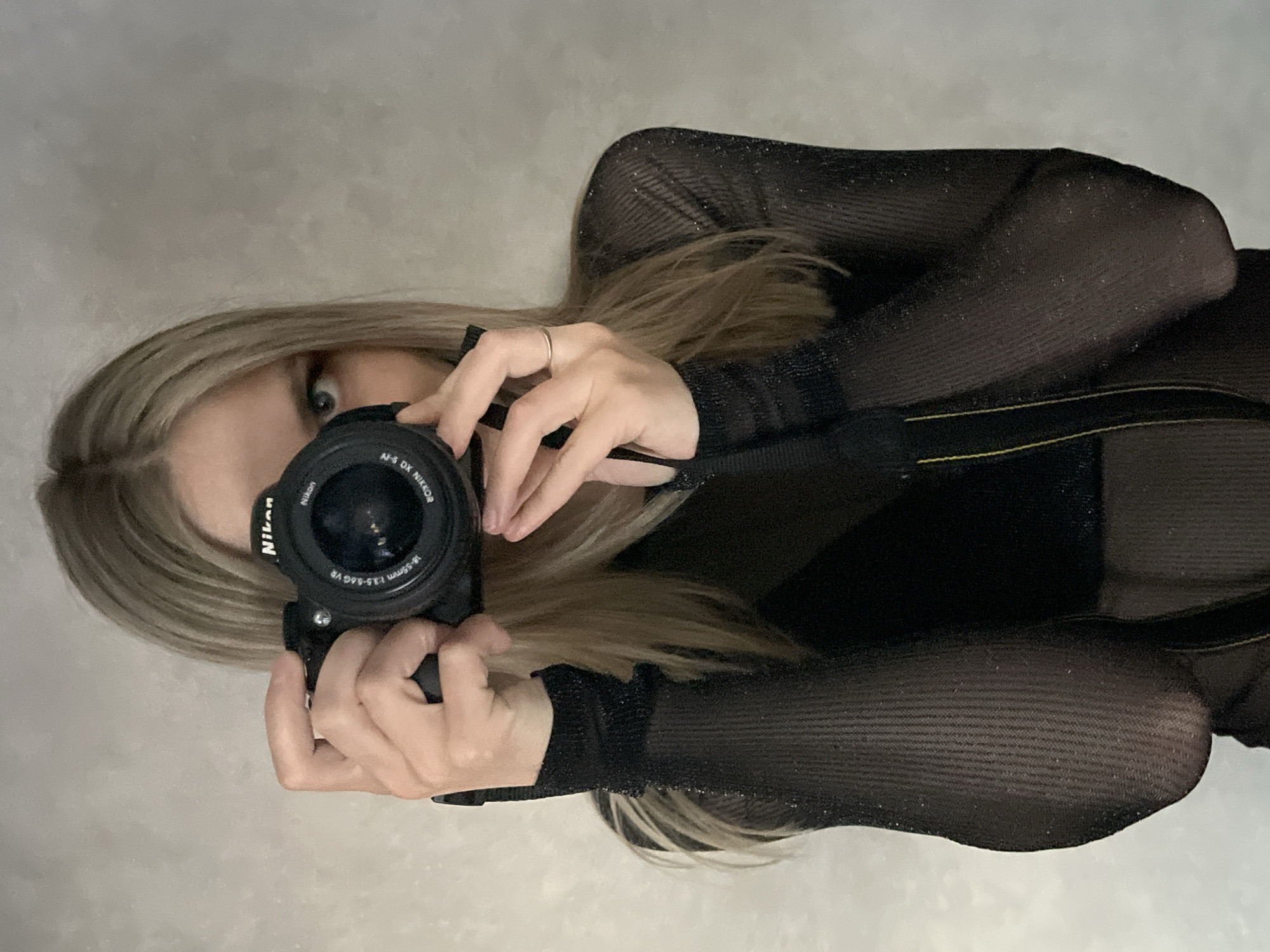 An image, originally oriented 90 degrees clockwise, depicts a person taking a photograph of themselves, likely in a mirror. The subject, a person with long, straight, light blonde or ash blonde hair, is positioned centrally. They are holding a black Nikon digital camera, which covers most of their face. One eye, appearing light-colored and wide open, is visible peeking over the top of the camera.
The person's hands are wrapped around the camera and lens. A simple silver band ring is visible on the ring finger of their right hand (from the subject's perspective). They are wearing a dark, possibly black, long-sleeved top made of a sheer or mesh fabric with a subtle glitter or shimmer effect. The fabric on the forearms has a ribbed texture. A dark strap, possibly from the camera or clothing, is also visible.
The camera is a Nikon model with "Nikon" visible on the body. The lens clearly shows the text "AF-S DX NIKKOR 18-55mm 1:3.5-5.6G VR". The background is a plain, light grey, subtly textured wall or surface, suggesting an indoor setting. The lighting appears even and diffused. While the location is provided as Chaikovsky, Russia, no visual cues within the image confirm this. The overall impression is one of a casual, self-portrait moment.