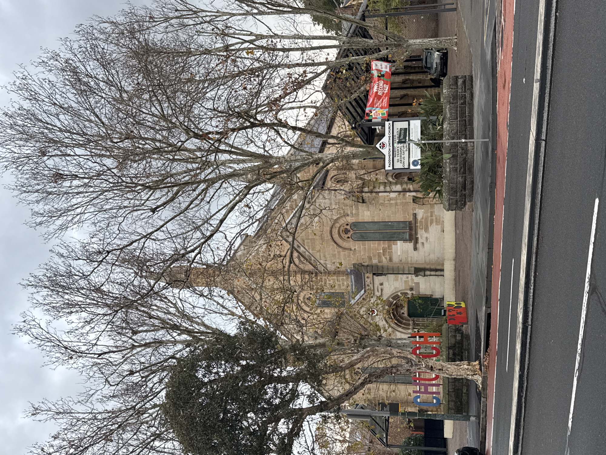 The image shows a stone church with a steeple, located in Sydney, Australia. In front of the church, there is a large sign that reads "PADDINGTON UNITING CHURCH". A red and white banner with the words "VINYL RECORD FAIR" is hanging to the right of the sign. In the foreground, there is a street with a red bike lane. Large bare trees with thin branches frame the church. A colorful sculpture of the word "CHURCH" is visible to the left of the entrance. The sky is overcast.