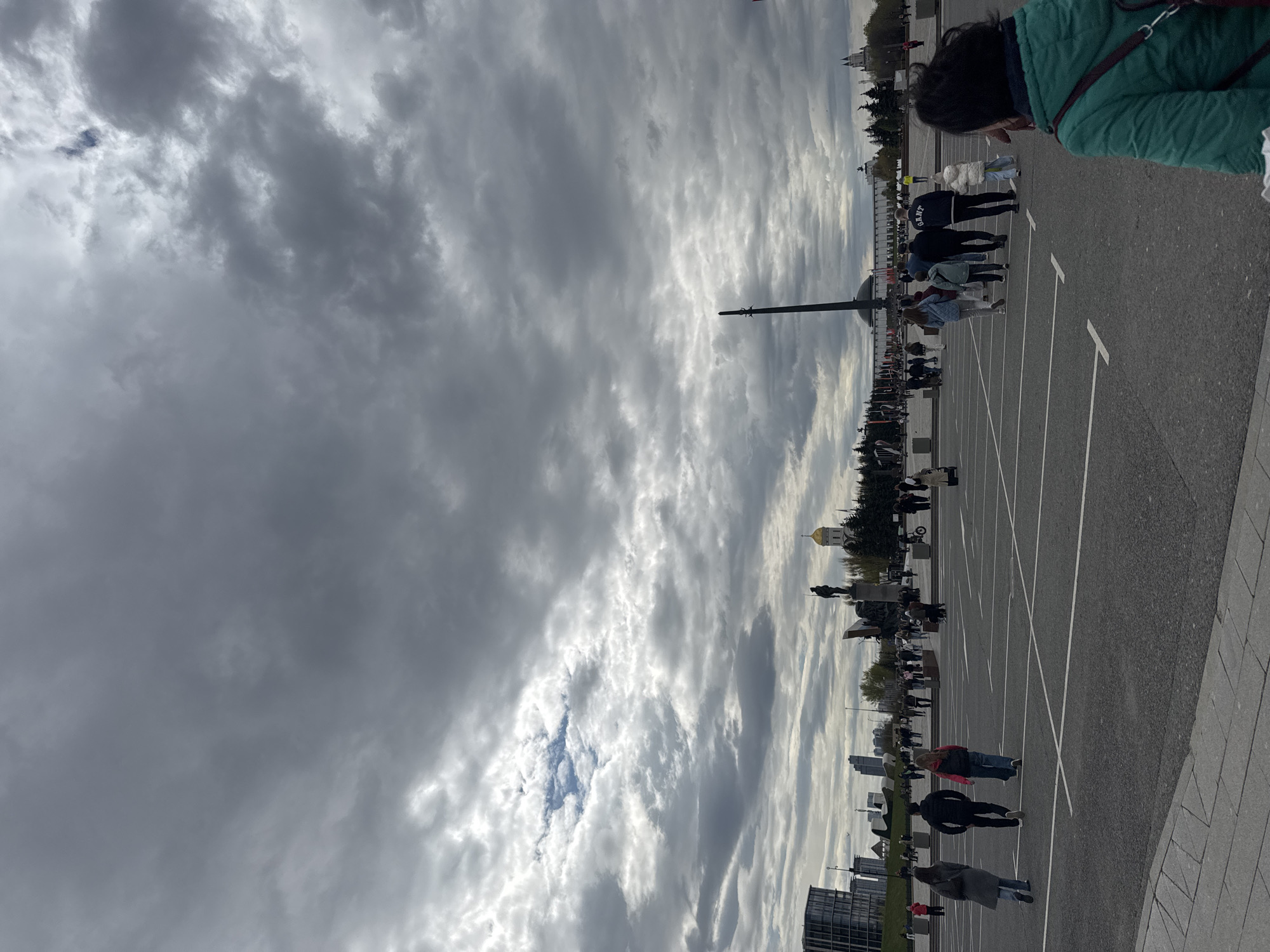The image shows a view in Moscow, Russia, likely a parking area near a monument. Above, there is a sky filled with layered clouds. On the right is the edge of a gray paved area with white parking lines stretching towards the background. People are scattered along the right side, mostly appearing as if they are walking or standing. Structures in the background include what appears to be a tall monument or obelisk, trees, statues, and buildings. The scene appears to be outdoors.