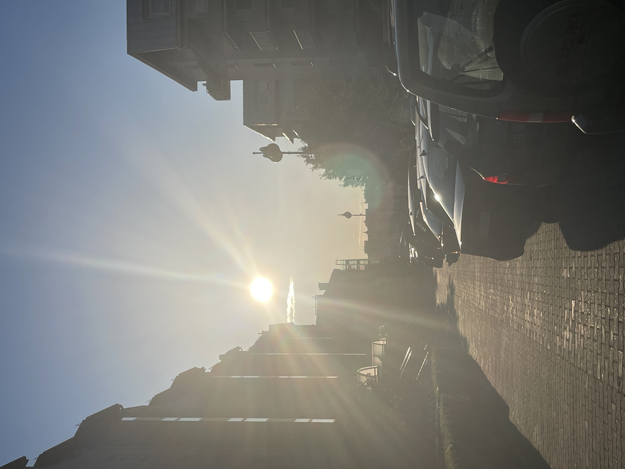 This image, set in an unknown city and country, depicts an urban street scene under strong, low-angle sunlight. The sun, located in the upper-left quadrant, generates intense luminosity, lens flare, and atmospheric haze across the frame. Multi-story buildings with pitched roofs are silhouetted along the left side, their windows reflecting the bright light. A cobblestone or brick pathway, bordered by a low hedge, extends along the left into the distance. A line of parked cars, silhouetted against the sun, occupies the right side of the street. The vehicle nearest the foreground on the right is an SUV with an external spare tire mounted on its rear. Further into the background, additional buildings and two distinctively shaped, elevated structures, resembling stylized streetlights or decorative poles, are visible. A small, horizontally oriented cloud is brightly illuminated directly above and slightly to the right of the sun. No people or overt actions are present.