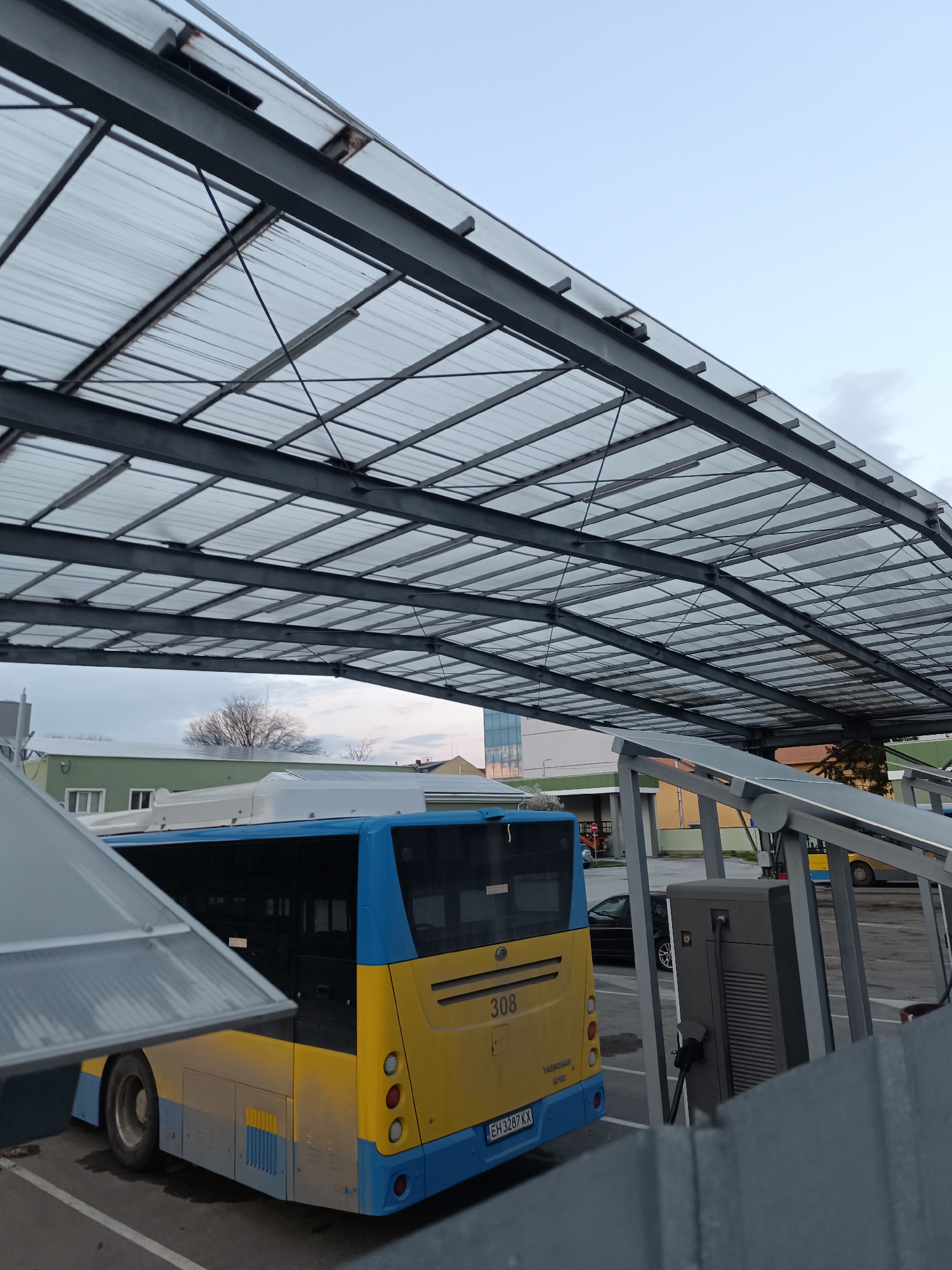 The image depicts a yellow and blue bus parked under a large, grid-like metal canopy structure. The bus, partially obscured by a foreground structure, has "308" visible on its rear and the license plate "EH3287KX". Next to the bus is an electrical charging station with a charging cable attached to the bus. The setting appears to be an urban charging station with other vehicles parked nearby. A building is visible in the background. The image was taken during overcast weather. The location is Pleven, Bulgaria.