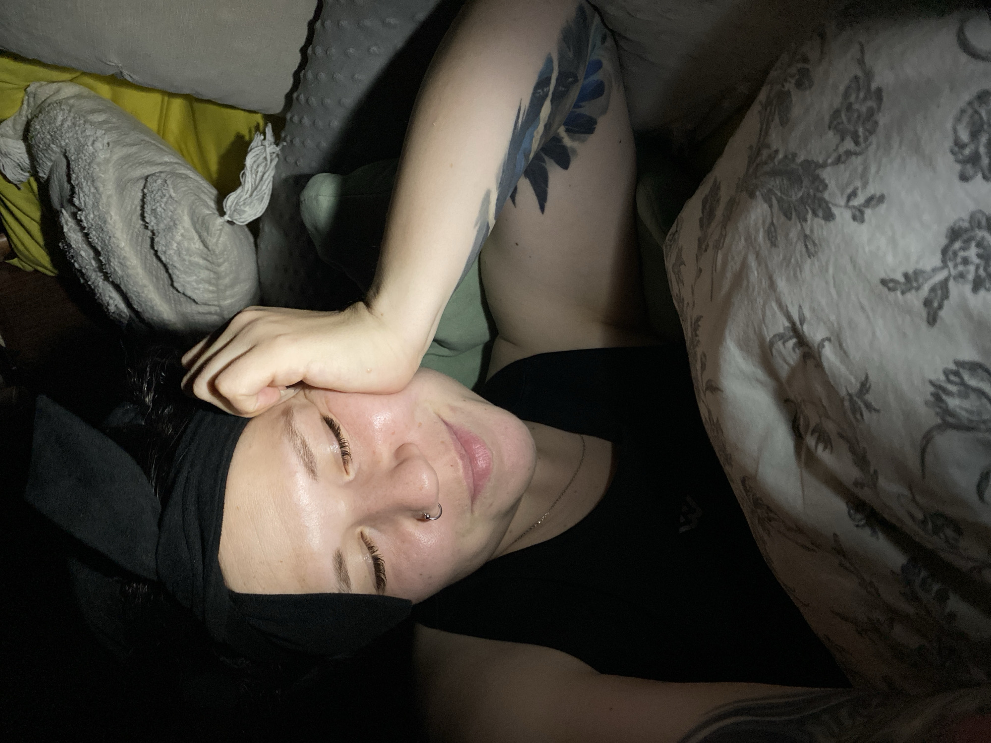 A woman is lying in bed. She is wearing a black headband and a black shirt. Her eyes are closed. Her left hand rests on her forehead. A tattoo is visible on her right arm. She is surrounded by pillows and a patterned duvet cover. The exact location in Chaikovsky, Russia, cannot be determined from the image.