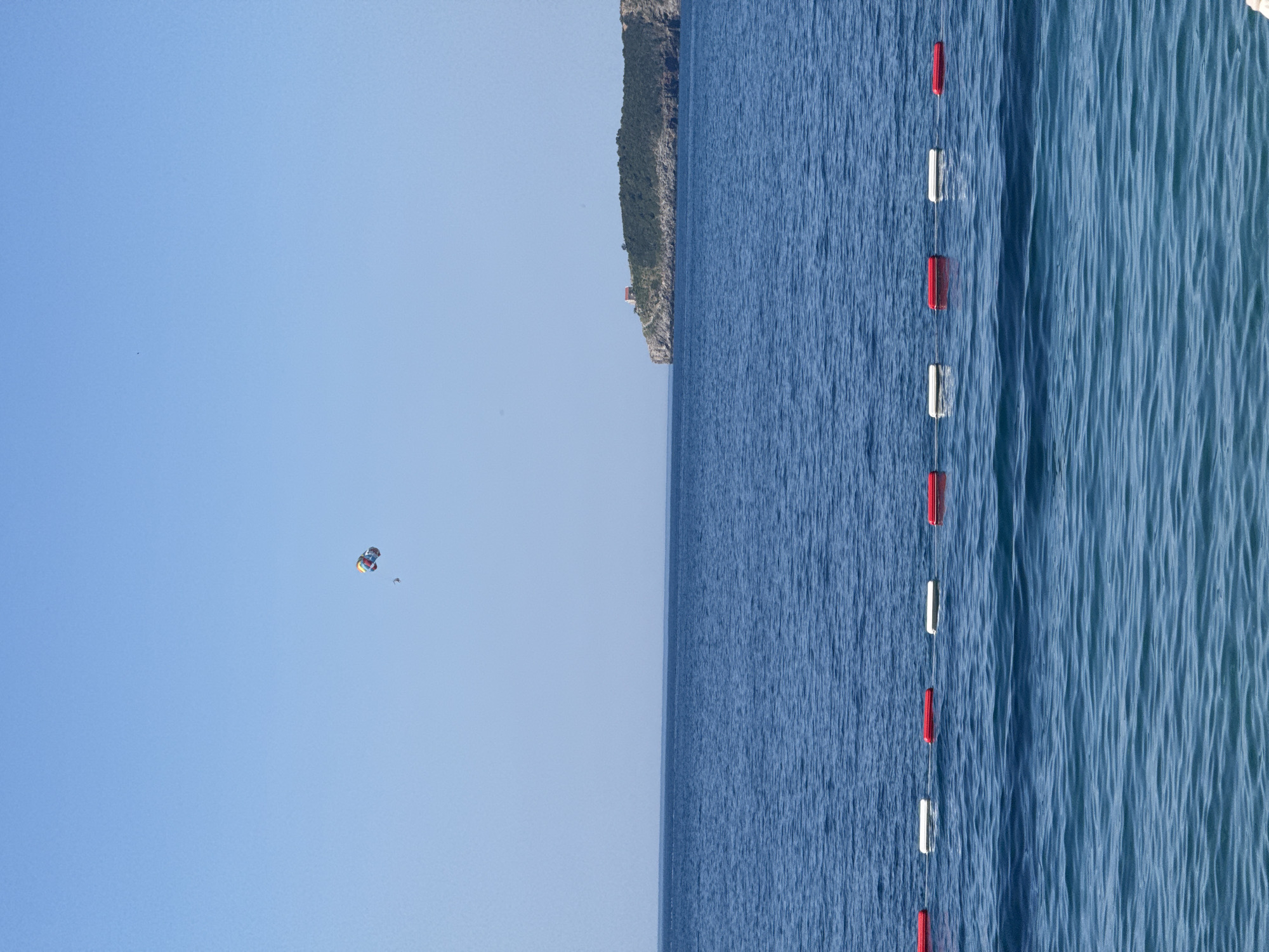 A parasailer is visible in the bright blue sky above a calm, deep blue sea. In the distance, a small island with a red-roofed building is situated on a rocky, tree-covered promontory. Along the foreground of the sea, a line of alternating red and white buoys separates the water, indicating a boundary for swimming or water sports. The image captures a serene coastal scene with elements of recreation and distant natural beauty.