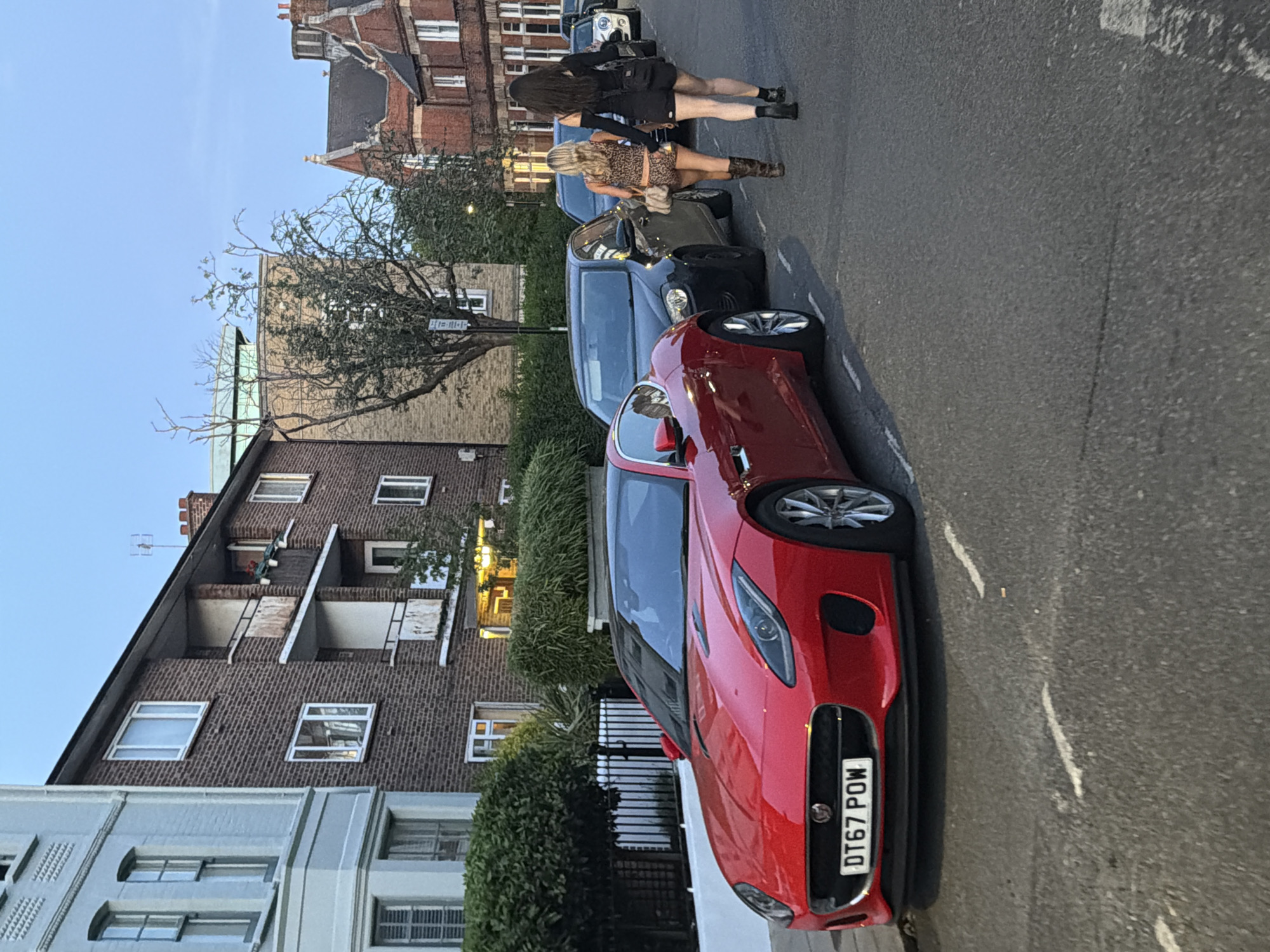 The image shows a street scene in London, United Kingdom. A red Jaguar sports car with the license plate DT67 POW is parked on the side of the road. Behind the Jaguar is a dark-colored sedan, followed by a lighter-colored sedan. Two women are walking on the side of the road next to the cars. One woman has long dark hair and is wearing a black dress and boots. The other woman has blonde hair and is wearing a leopard print top and brown boots. Buildings line the street, including brick apartment buildings and a light-colored building with multiple windows. A tree is present between the buildings and the cars. The sky is light blue, suggesting it's late afternoon or early evening. The street is asphalt with painted white lines marking parking spaces.