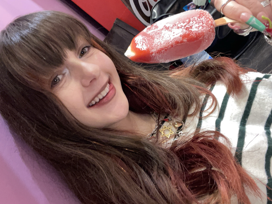 A close-up selfie captures a person, likely a young woman, smiling brightly while holding a red frozen treat. She has long, dark brown hair with bangs that fall across her forehead, and visible reddish or auburn streaks/highlights throughout her hair, particularly on the right side. Her eyes are light brown, and she has a wide, happy smile, showing her teeth. Her tongue is slightly visible, playfully touching her upper lip.
She is holding a red, elongated frozen treat, possibly a popsicle or ice cream bar, on a wooden stick in her right hand. The treat is covered in a layer of white frost, indicating it's very cold. Her nails are long and manicured, appearing to have a light green or bluish color with some additional design or glitter. Several rings are visible on her fingers.
Around her neck, she wears a necklace with multiple charms, prominently featuring a large, golden Star of David pendant in the center, flanked by other smaller charms. She is wearing a white garment with dark green or black horizontal stripes, part of which is visible over her right shoulder and chest area.
The background on the left side of the image is a solid, light pink or purple color. On the right, the background is darker, with a blurry red and black object or pattern, possibly a logo or sign, but it is not clearly decipherable. The image is taken at a slight upward and angled perspective, suggesting a selfie or a casual close-up shot. The overall mood conveyed is cheerful and relaxed.