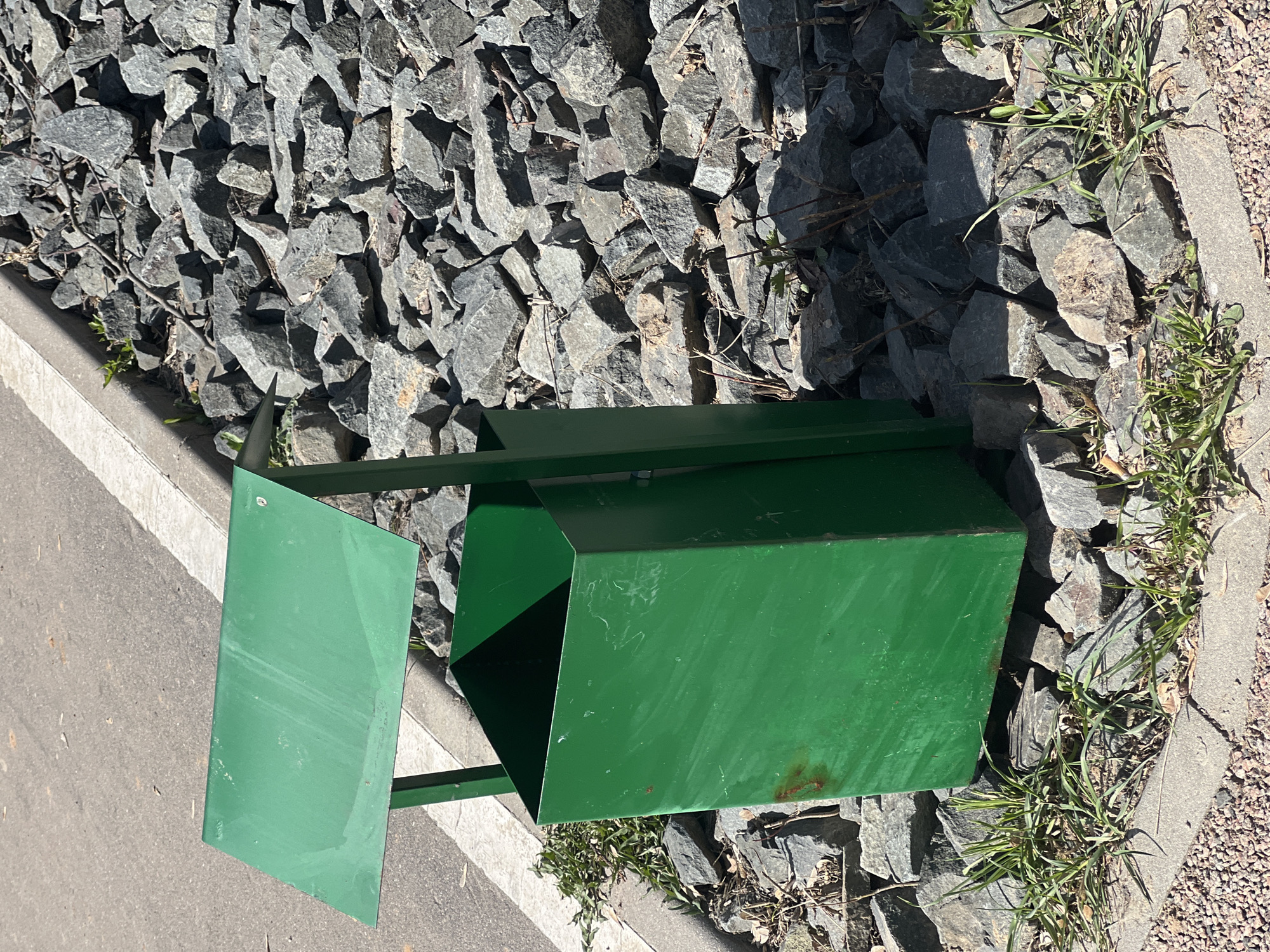 A green metal trash receptacle with a hinged door stands on a bed of grey stones next to a paved path in Khimki, Russia. The trash can is positioned between the path and a concrete curb with grass growing near it.