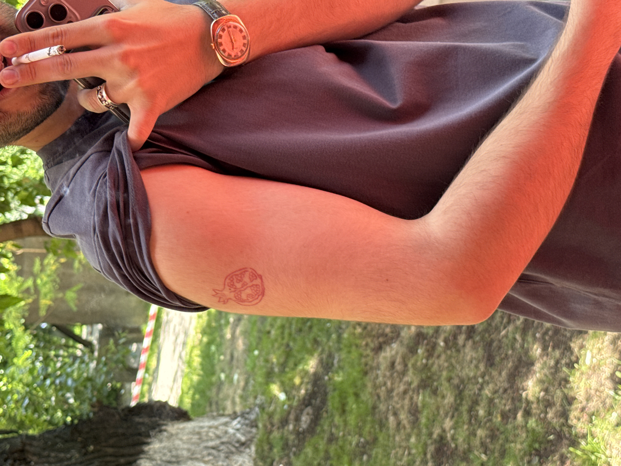 The image shows a close-up of a person's upper right arm, on which a small, minimalist tattoo of a pomegranate is visible. The person is wearing a dark gray t-shirt and a watch with a brown leather strap. They are holding a cigarette in their right hand and a smartphone to their ear. The background suggests an outdoor setting, possibly a park or garden, with greenery and trees. A red and white striped ribbon is visible in the background, suggesting a temporary barrier or boundary. The lighting indicates it is daytime, with a warm, reddish hue cast over the scene, possibly due to the time of day or artificial lighting. The location is stated to be Yerevan, Armenia.