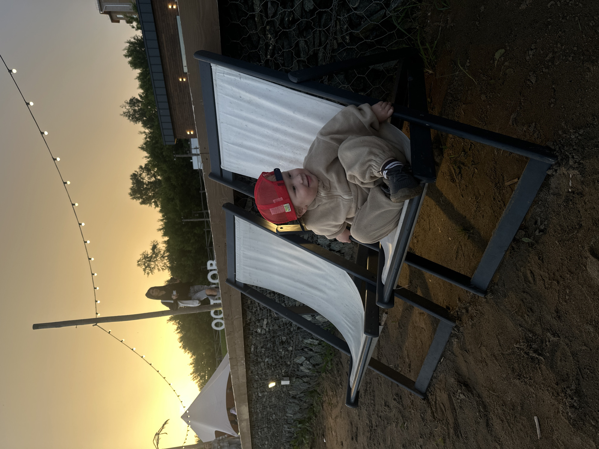 An outdoor scene at sunset features a young child sitting on a white deck chair. The child, wearing a tan hooded jumpsuit and a red baseball cap, is looking to the right with a slight smile. The deck chair is one of two visible, both with black frames and white canvas seats, positioned on a sandy surface. In the background, string lights with bulbs are suspended across the frame. Further back, a wooden structure with dark siding is visible, and beyond that, a line of trees. Two other figures, a woman and a child, are partially visible in the distance, near a white tent-like structure. The overall ambiance suggests a recreational area. The location is identified as Votkinsk, Russia.