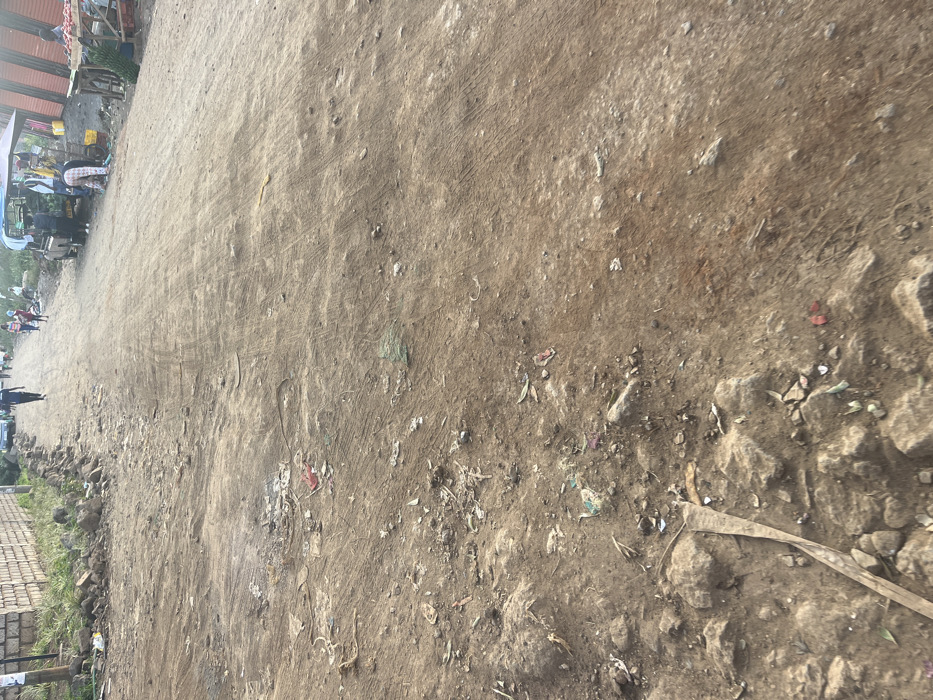 The image displays an unpaved dirt road in Gitothua ward, Kenya, viewed from a low-angle perspective. The foreground features an uneven, compacted earth surface strewn with rocks, small stones, and scattered debris, including plastic remnants and organic material. Visible tire tracks indicate vehicular passage. Along the left margin of the road, a low stone wall is visible, accompanied by a loose pile of larger stones and sparse vegetation. Further along this side, a blue sedan is parked. Several individuals are present on the road: one person in dark attire walks away from the viewer, and other figures are distributed further into the background. On the right side of the road, multiple makeshift market stalls are active. One stall includes a light-colored canopy positioned over a utility vehicle or display area, where an individual is bending over, interacting with merchandise. Adjacent to it, another stall, constructed from corrugated metal sheeting, showcases produce on a wooden table. The scene depicts a functional, active commercial thoroughfare with pedestrian and vehicular movement.