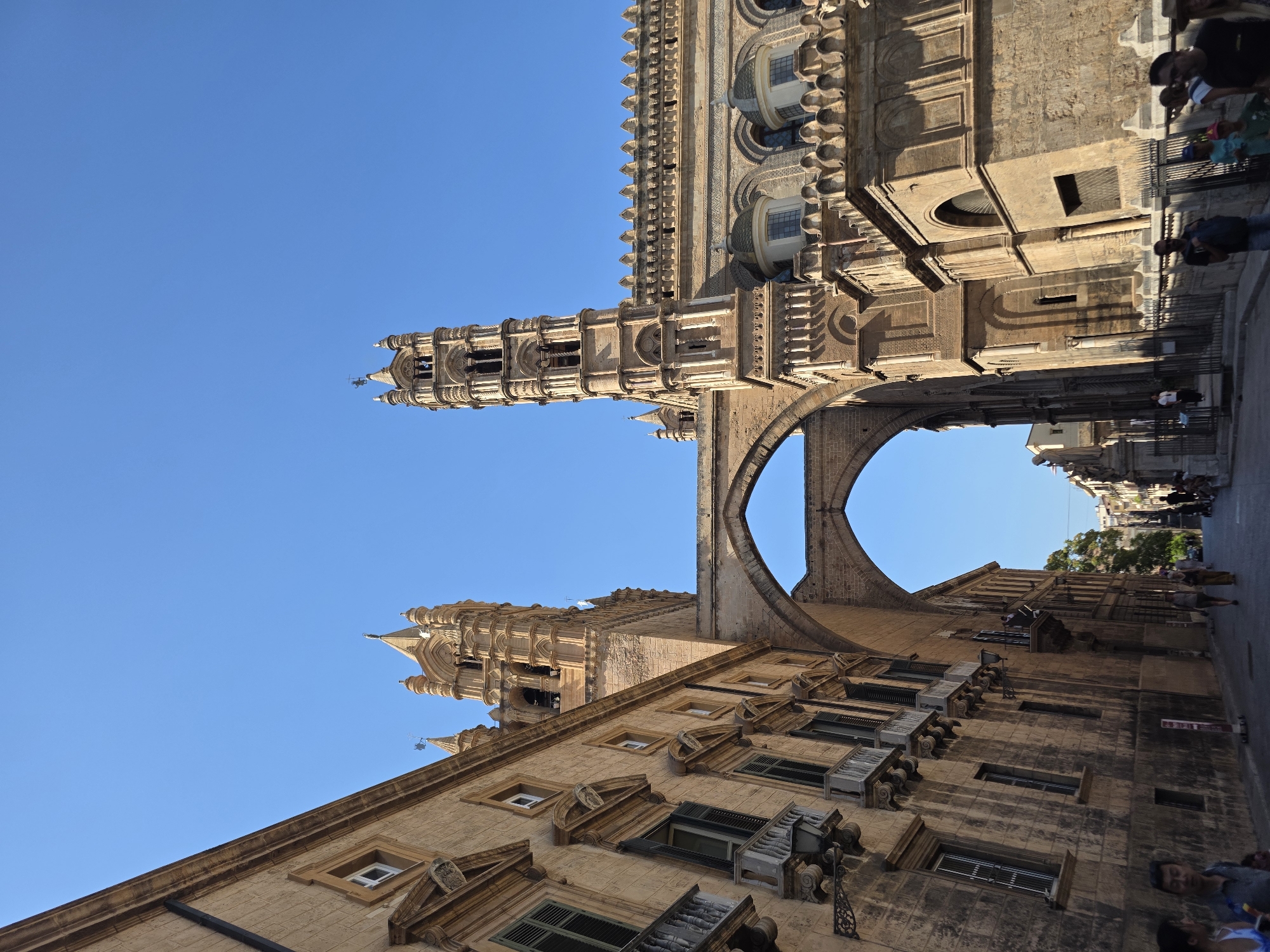 The image shows a low-angle view of the exterior of a large, ornate building with a clear blue sky above. The architecture is a blend of styles, featuring tall, intricately carved towers with pointed spires, and a large arched structure connecting two parts of the building. The main structure is made of light-colored stone, with decorative elements such as arched windows, small domes, and patterned stonework along the roofline. To the left of the frame, another building with similar architectural features, including balconies and shuttered windows, is visible. A street runs in front of these buildings, with several people walking along it. Some individuals are in the foreground, while others are further down the street. The overall scene suggests an urban environment, likely a historic city center.