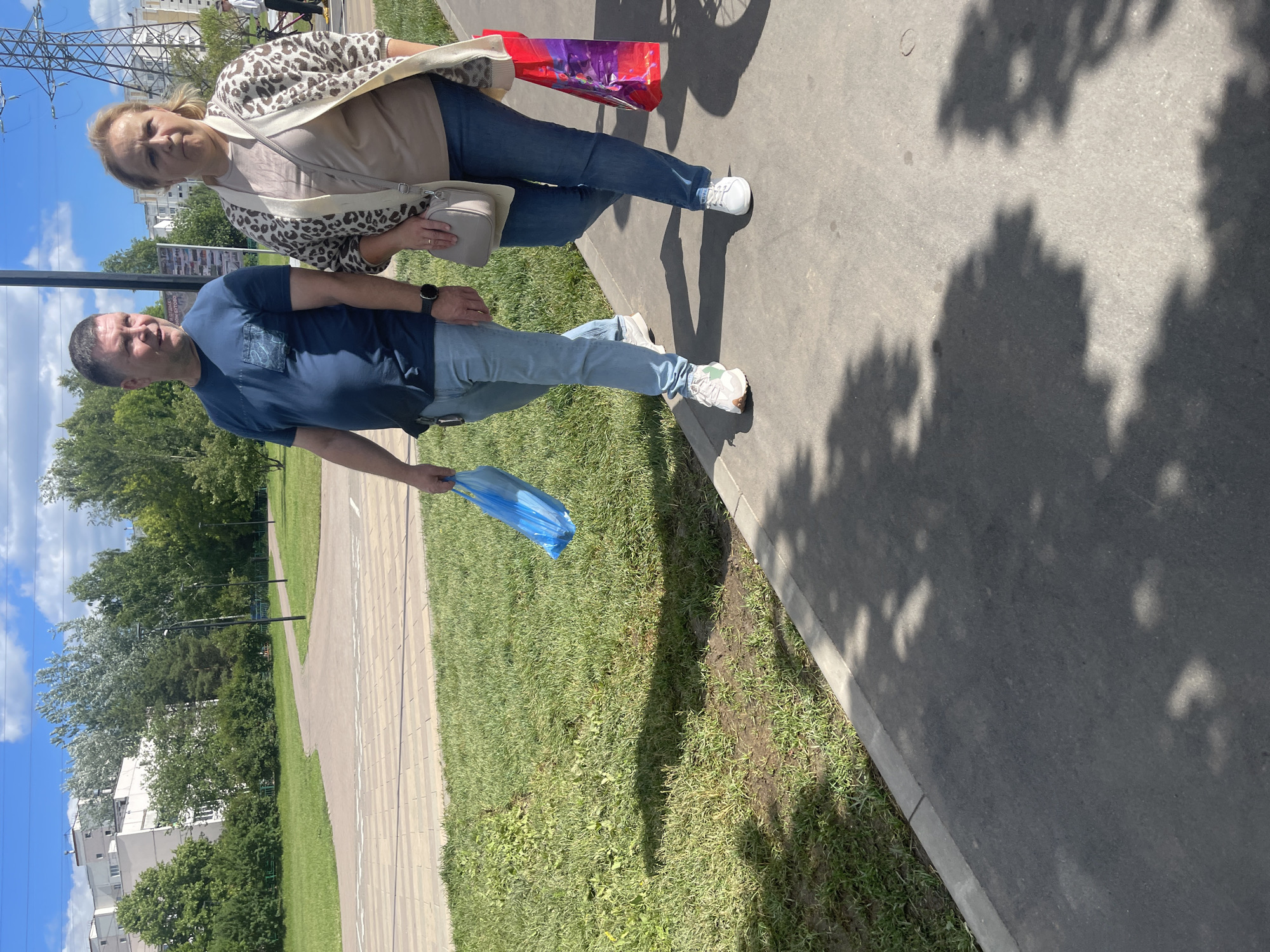 The image shows two people, a man and a woman, walking on a paved path next to a grassy area. The man, in the foreground, is wearing a dark blue t-shirt and light blue jeans. He is holding a blue plastic bag in his right hand. The woman, in the background, is wearing a beige shirt and a leopard-print cardigan over it, also in light blue jeans. She is carrying a light-colored handbag. They are walking away from the camera.
The setting appears to be a park or a residential area with trees and a building visible in the background. The sky is blue with some clouds, suggesting it is daytime and likely fair weather. The shadows cast on the path indicate the sun is shining. The presence of a power line tower in the distance and the architectural style of the building might suggest an urban or suburban environment in Moscow, Russia. The overall mood seems casual and everyday, with no indication of a specific event. There is no visible text in the image.