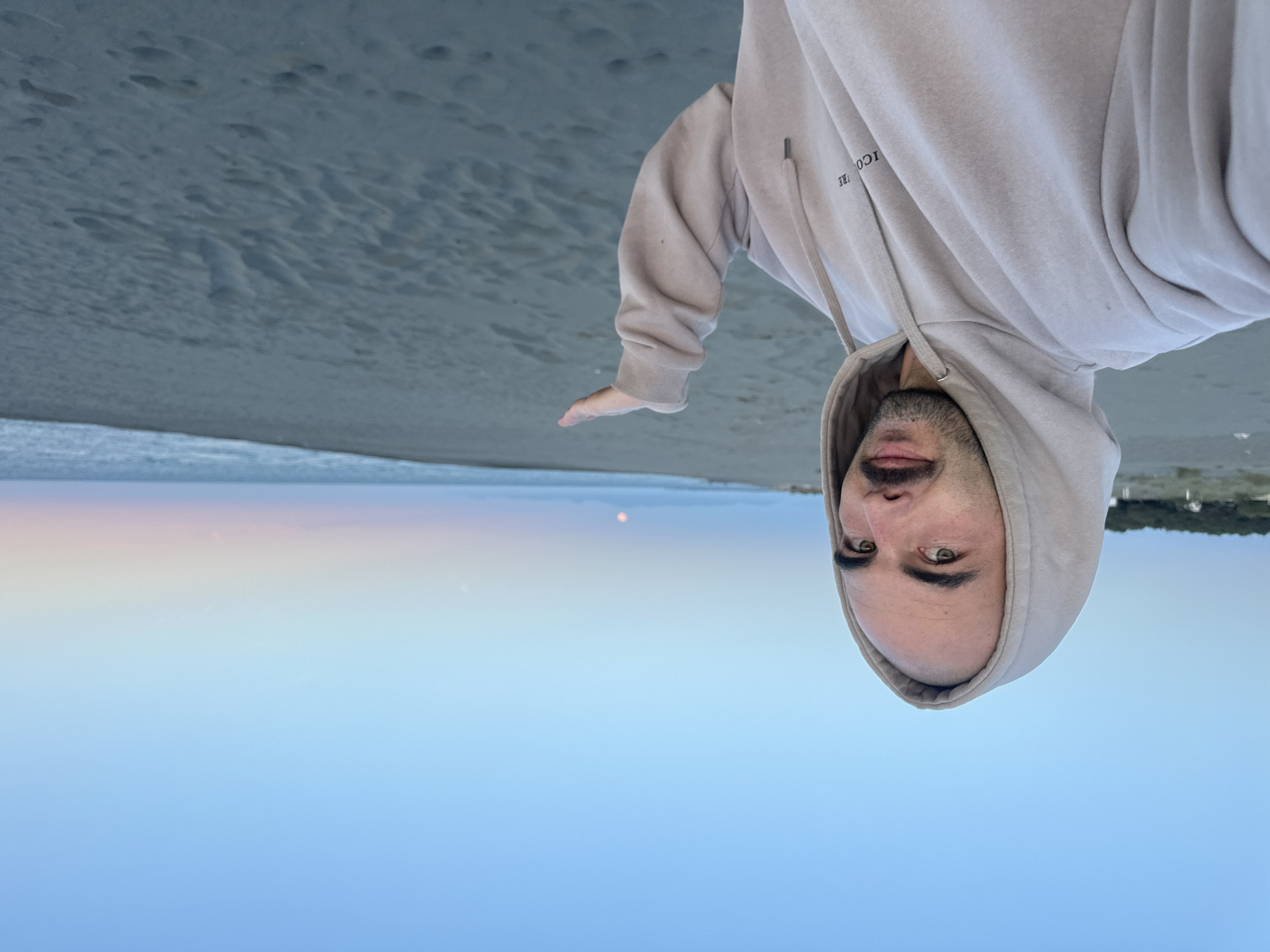 A man wearing a hooded sweatshirt is standing on a beach. The background shows the sea, the sky, and a distant landscape. He is likely at Velika Plaža in Donji Štoj, Montenegro.