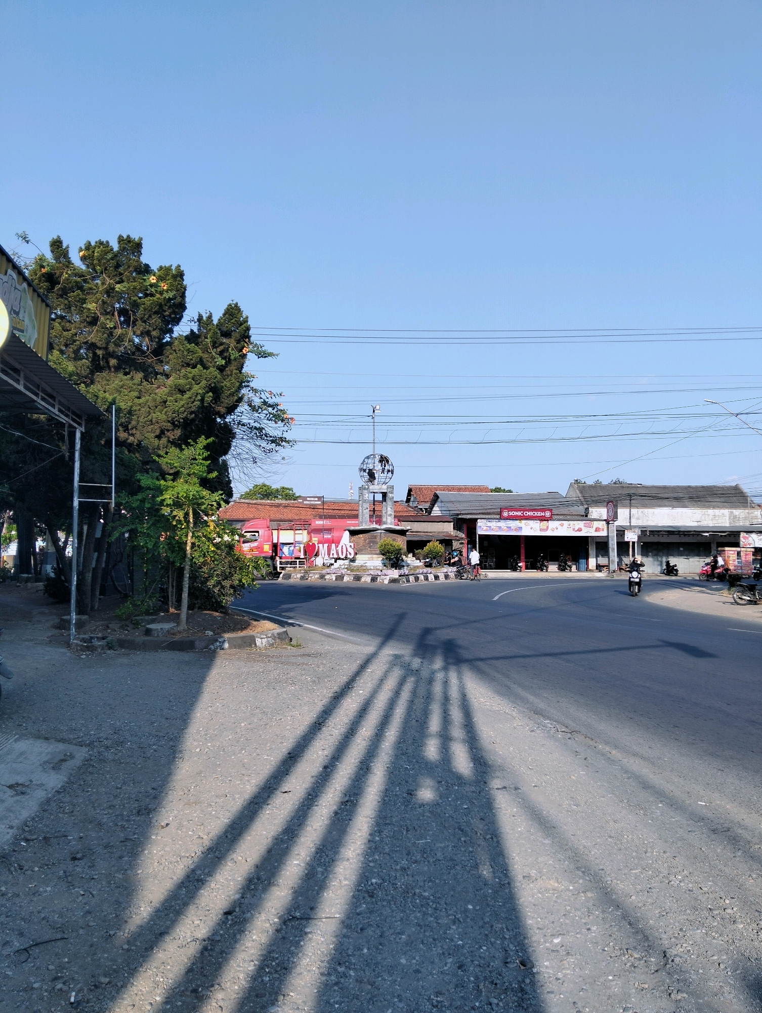 The image depicts a street scene in Desa Karangkemiri, Indonesia. In the foreground, a paved road is visible, with distinct shadows cast across it, indicating a bright sunny day. To the left of the road, a sidewalk area is present, bordered by vegetation including trees and bushes. Beyond the vegetation, a red truck is parked. In the middle ground, there are several commercial buildings lining the street. One building prominently displays a "SONIC CHICKEN" sign. Further down the street, a roundabout is present, with a globe-like monument at its center. Several motorcycles are parked and some are in motion on the road, suggesting moderate local activity. The sky is clear and blue. The overall environment appears to be a commercial area within a town or village, possibly near a traffic intersection or monument.
