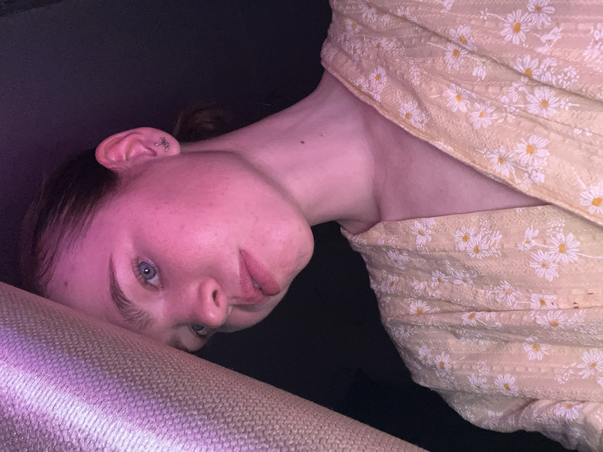 A close-up, angled portrait shows a fair-skinned person with light blue eyes, resting their head on a beige, textured surface, possibly a cushion or sofa. The person is mostly bathed in pinkish light, with their face tilted towards the camera. They have reddish-pink lips, visible eyelashes and eyebrows, and their hair is pulled back. An ear is visible, adorned with a silver-colored piercing. The person is wearing a yellow floral-patterned garment or top. The background is dark. There is no indication of this photo being taken in the city of Chaikovsky, Russia.
