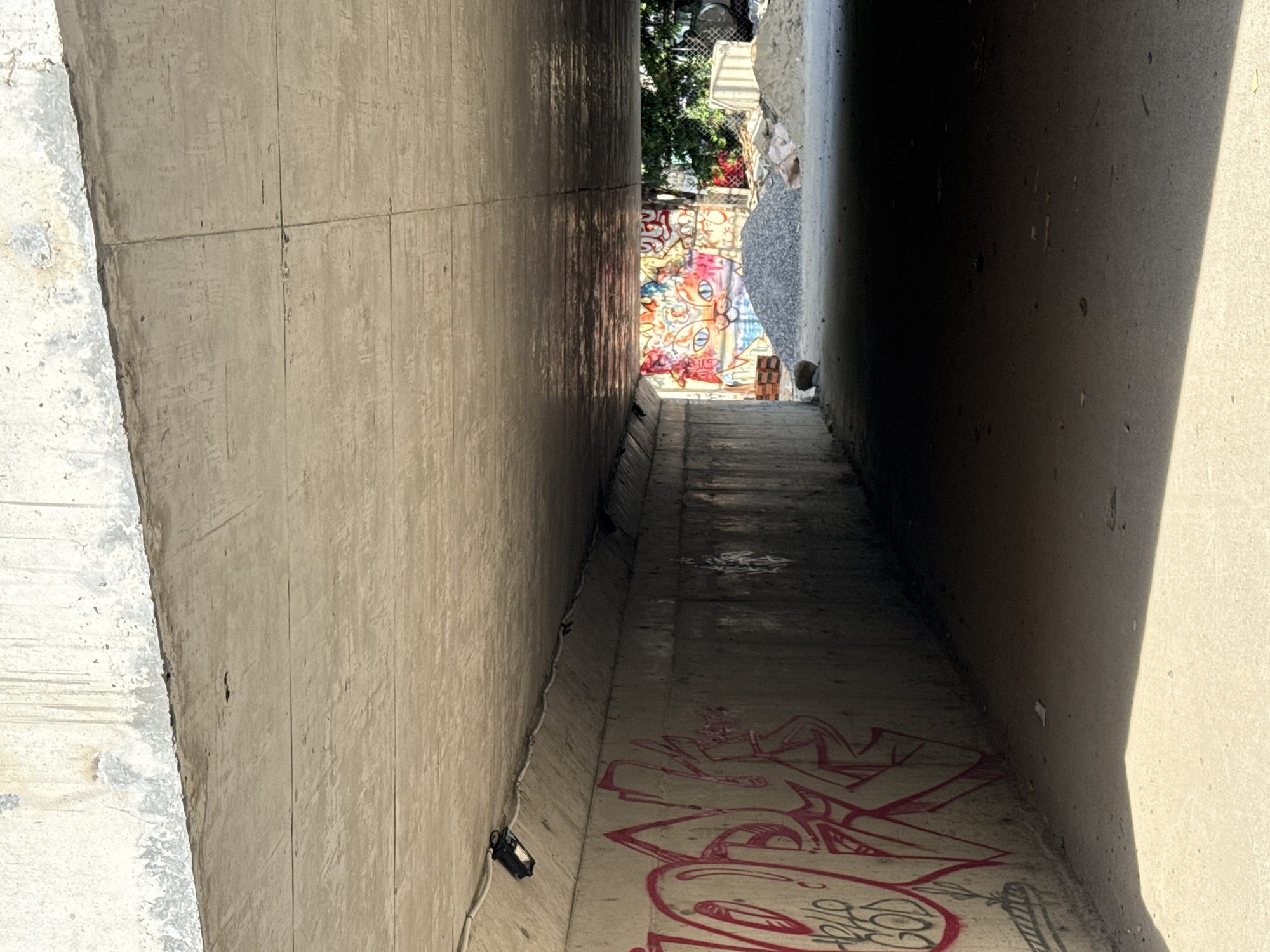 The image shows the interior of a concrete underpass or tunnel. The ceiling is made of rough concrete, showing signs of wear and tear, with some water stains. The walls of the underpass are also concrete, with one side covered in vibrant graffiti. The graffiti on the left features large, stylized red lettering and some smaller text that appears to read "HELLO GOD." The right wall recedes into shadow.
At the end of the underpass, a brightly colored mural of a cat's face is visible, painted on a wall. In front of the mural, there are piles of gravel and sand, suggesting construction or maintenance work is taking place. The ground within the underpass is paved with concrete and shows some scattered debris. Sunlight streams in from the end of the tunnel, creating distinct areas of light and shadow. The overall environment appears to be an urban underpass, likely in an area undergoing development or renovation. The specific location in Bac Nha Trang Ward, Vietnam, is inferred from the prompt but not visually evident within the image itself. There are no people visible in the scene.