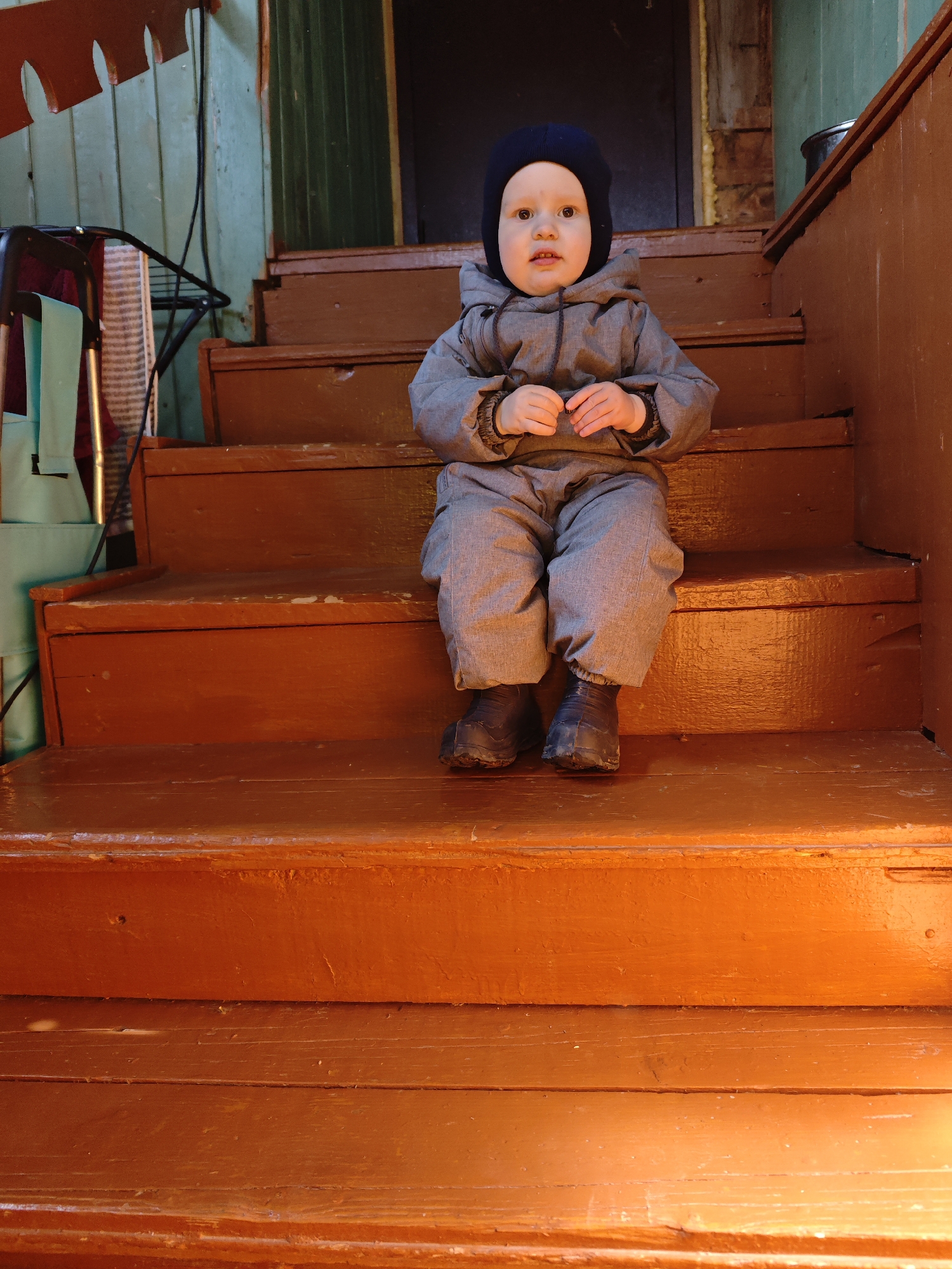 A young child, likely a toddler, is depicted sitting on a wooden staircase in Фоки, Russia. The child is positioned on the third step from the bottom, looking directly towards the viewer with a calm, wide-eyed expression.
The child is dressed in warm attire, indicating cool or cold weather. They wear a dark blue balaclava or beanie that covers their head and ears, and a grey, possibly insulated or waterproof jumpsuit. The suit features elasticated cuffs on the wrists and ankles, and a zipper down the front. Their feet are encased in dark, practical boots.
The setting appears to be an outdoor porch or an unheated entrance area of a building. The stairs are made of wood and are painted a reddish-brown or warm orange color, showing some signs of wear and age. In the background to the left, there is a metallic frame structure, possibly part of a stroller or wheeled cart, with a light teal fabric bag or cover hanging from it, and a striped fabric item beside it. The wall behind the child is painted a pale green, consisting of horizontal wooden planks. A dark doorway or opening is visible further back. A black electrical wire runs vertically on the left side of the frame. The lighting suggests it is daytime, with soft, diffused light. There is no visible text in the image.