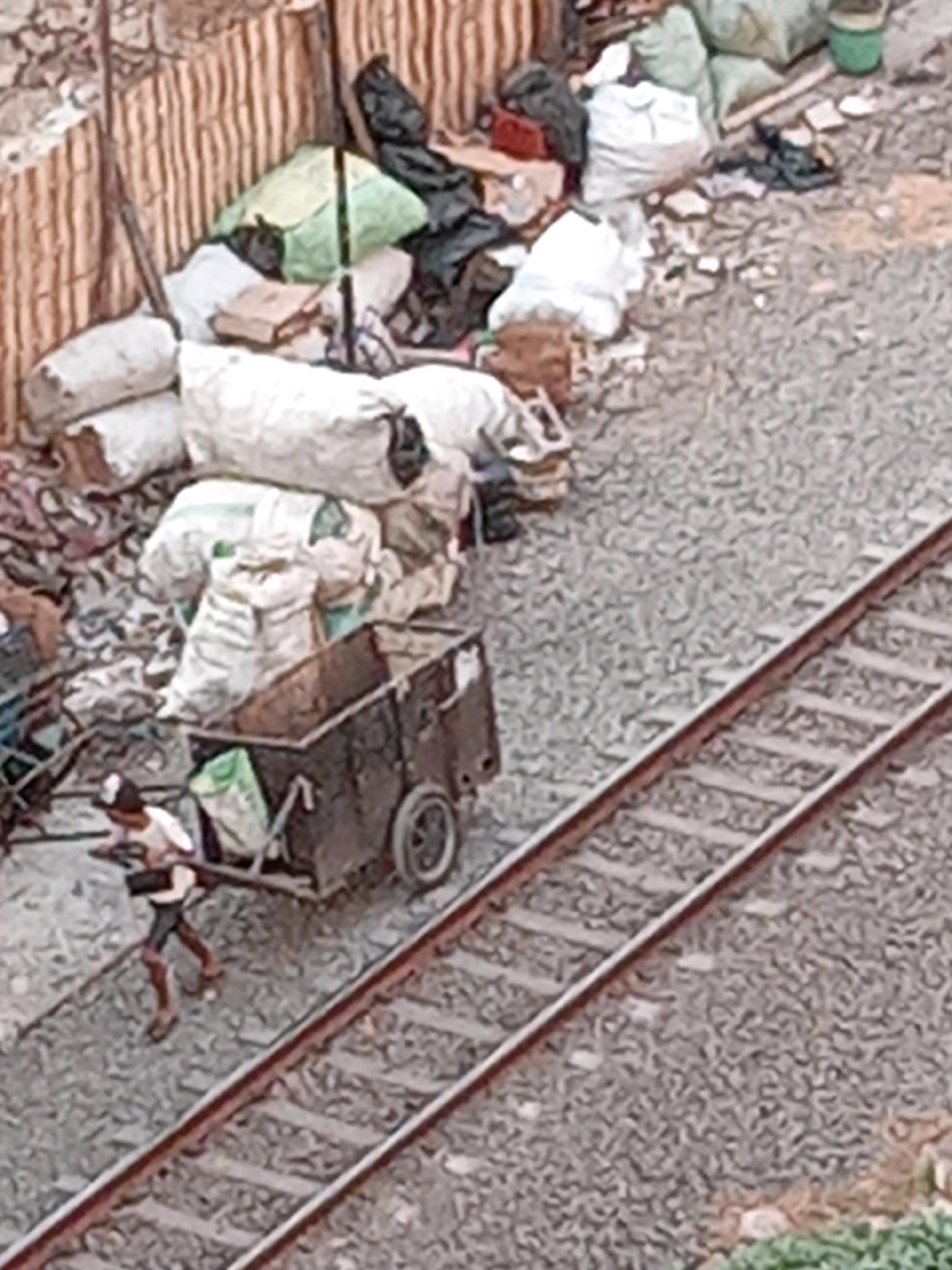 A person is pushing a cart filled with sacks alongside a railway track in what appears to be a slum area. The cart is old and metallic, with two wheels, and it is loaded with several large, white sacks and one green sack. The person pushing the cart is wearing a white t-shirt and dark shorts, with their back to the camera. The railway track is made of gravel and steel rails. In the background, there is a pile of various sacks, some green and some white, along with other debris. A wooden fence-like structure is visible behind the pile of sacks. The overall environment suggests a neglected or informal settlement, likely within the city of Surabaya, Indonesia, given the context of the prompt. The scene depicts an everyday activity of transporting goods, possibly for recycling or sale, in a challenging environment.