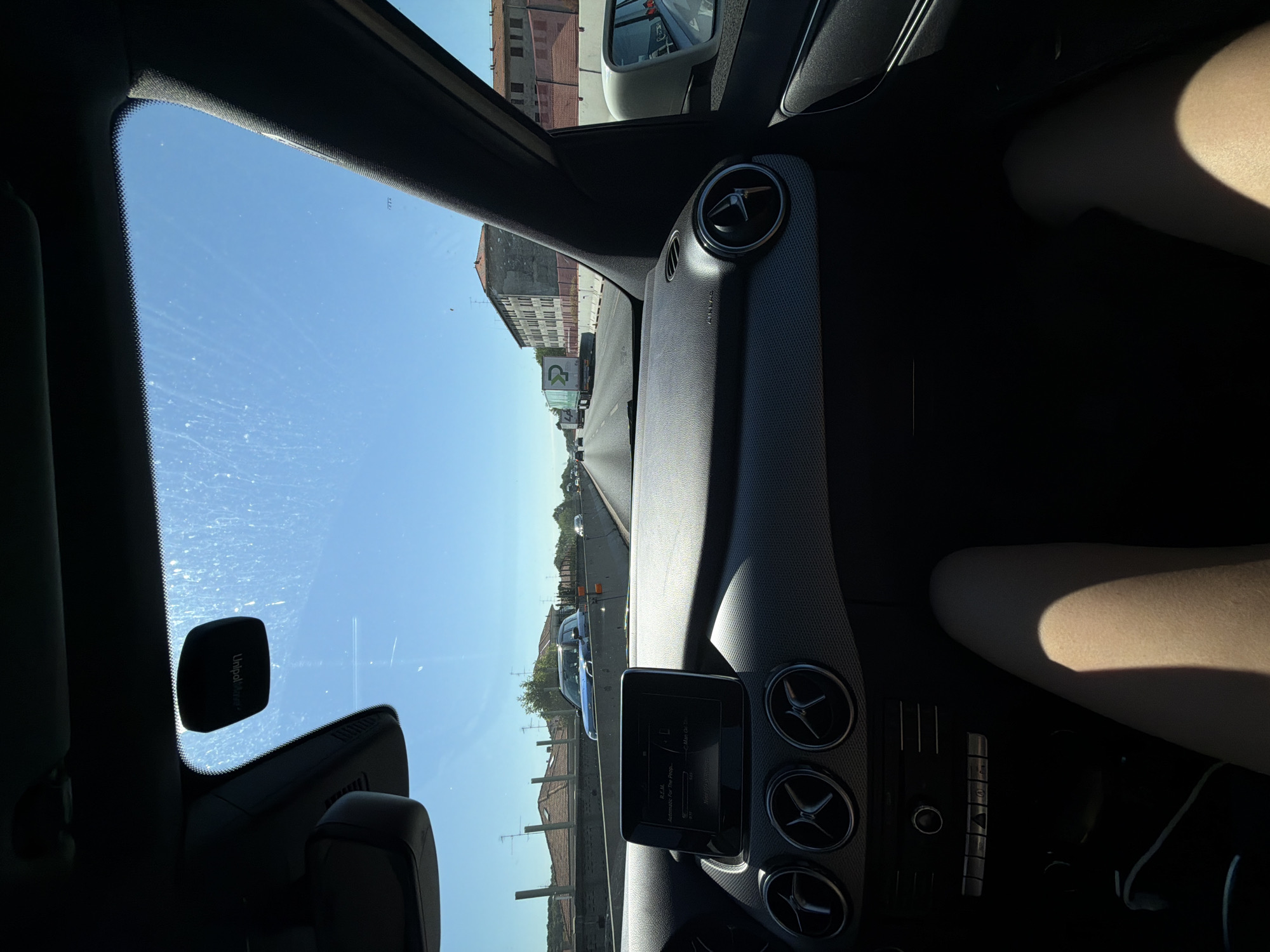 The image captures a view from inside a car, looking out through the windshield and side window. The car's interior is visible, including the dashboard with air vents and a central infotainment screen displaying track information for "Automobile For The Prophecy" by R.E.M. The perspective suggests the car is in motion on a highway.
Outside, the scene is dominated by a clear blue sky, indicating a sunny day, likely midday given the bright sunlight. There are buildings with tiled roofs visible along the side of the road, suggesting an urban or suburban environment. A truck with a green logo is seen in traffic ahead. The presence of other vehicles indicates that a normal traffic flow is occurring.
On the right side of the interior, a person's legs are visible, illuminated by sunlight casting a shadow. The person appears to be a passenger. The overall mood is one of travel on a pleasant day.
Visible text includes "AIRBAG" on the dashboard, "UnipolMoveR" on a device mounted on the windshield, and the song title and artist on the infotainment screen. The location context provided places the scene in Castellanza, Italy.