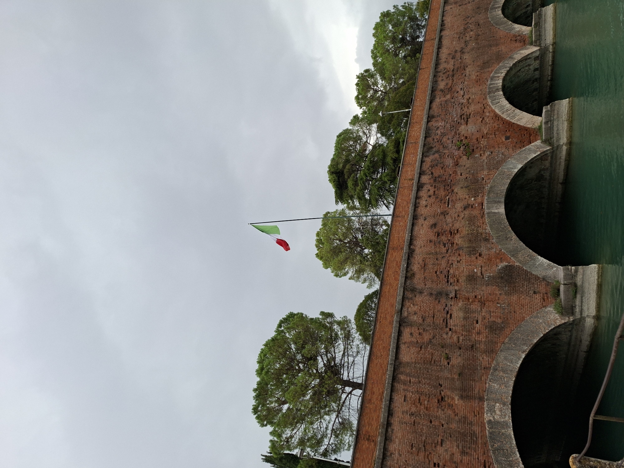 The image depicts a brick bridge with arched openings over green water. An Italian flag waves from the top of the bridge, which is lined with trees. The sky is overcast. The location is Peschiera del Garda, Italy.