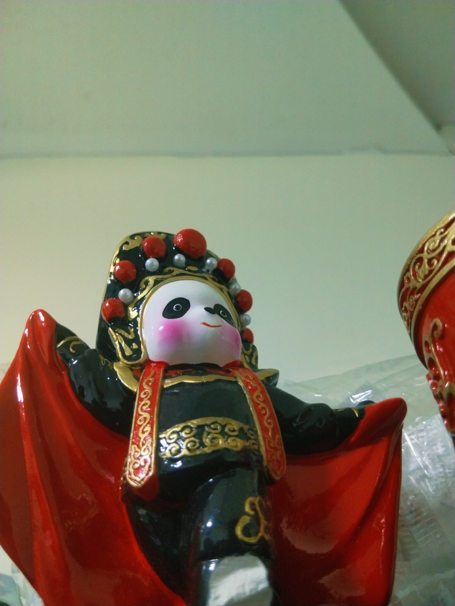 The image depicts a ceramic figurine of a panda dressed in the style of a Peking opera performer. The panda has a white face with black patches around the eyes, pink cheeks, and a small, smiling mouth. It wears a black and gold headdress adorned with red and pearl-like embellishments. The panda is dressed in a black and gold outfit with a red, ornate trim. A red cape-like garment extends from its shoulders. To the right of the panda, a portion of another similar figurine is visible, featuring red and gold patterns. The background is a plain, light-colored wall or surface. The location is Unknown, Unknown.