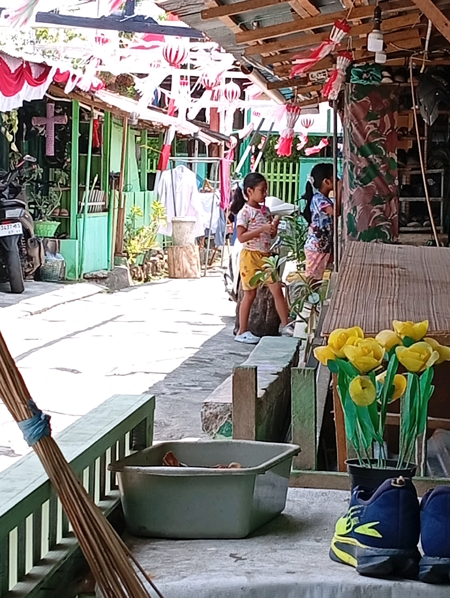 The image depicts a street scene in Manado, Indonesia, possibly in front of a residential property. Red and white decorations, resembling Indonesian flags and lanterns, are strung across the foreground and midground, suggesting a celebratory occasion. Two young girls, one in a pink and white patterned top and yellow shorts, the other in a blue and white patterned shirt and pink shorts, are visible in the midground. They are standing near a wooden structure and appear to be interacting with something in their hands. In the foreground, a broom is leaning against a green wooden railing. To the right of the broom, a large grey basin rests on a table-like surface. Further to the right, a potted plant with yellow flowers and a pair of blue and yellow sneakers are present. The background features a green building, a motorcycle, and more decorations, as well as shelves stocked with items. The lighting suggests it is daytime, with strong shadows cast on the ground, indicating the sun is high.