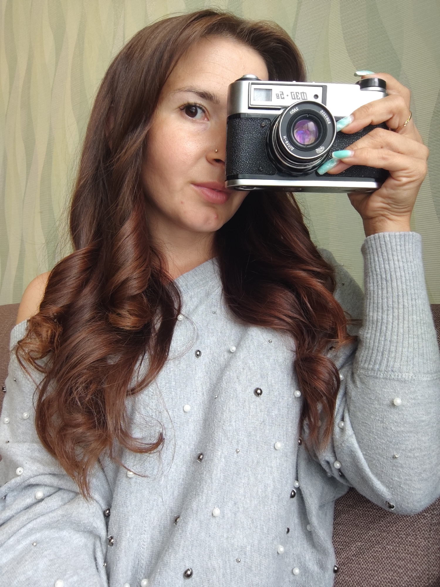 A woman with long, brown curly hair is holding a vintage camera in front of her face, partially obscuring her left eye. She is wearing a gray sweater adorned with pearl and silver-colored embellishments. Her nails are painted with a light blue marble-like pattern. A gold ring is visible on her right hand. The background is a textured wall of light green with subtle stripes. The location Фоки, Russia is not discernible from the visual data.