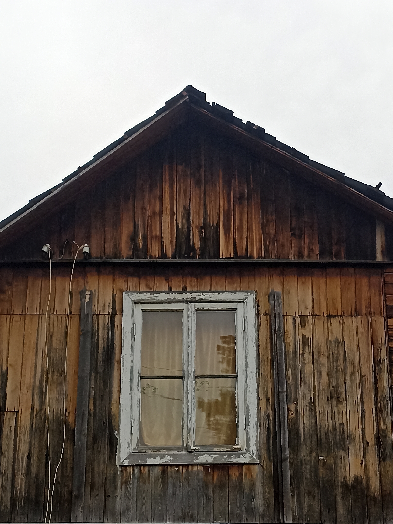The image shows a close-up, low-angle exterior view of an old, weathered wooden building. The building's facade is composed of vertical wooden planks of varying shades of brown and gray, indicating age and exposure to the elements. Some areas show dark staining, possibly from water damage or rot. The roofline is visible at the top, forming a sharp peak covered with dark shingles. A single window with a light-colored, peeling frame is centrally located in the middle section of the building. The window panes are somewhat obscured by what appears to be sheer, light-colored curtains. On the left side of the building, near the roofline, two white ceramic electrical insulators are mounted, with thin wires strung between them and extending downwards. The sky above is overcast and a uniform pale gray. The scene appears to be devoid of human activity or any immediate signs of life. The image does not provide any specific geographical markers to identify its location within Mochische, Russia, but it depicts a typical rustic or older rural structure.