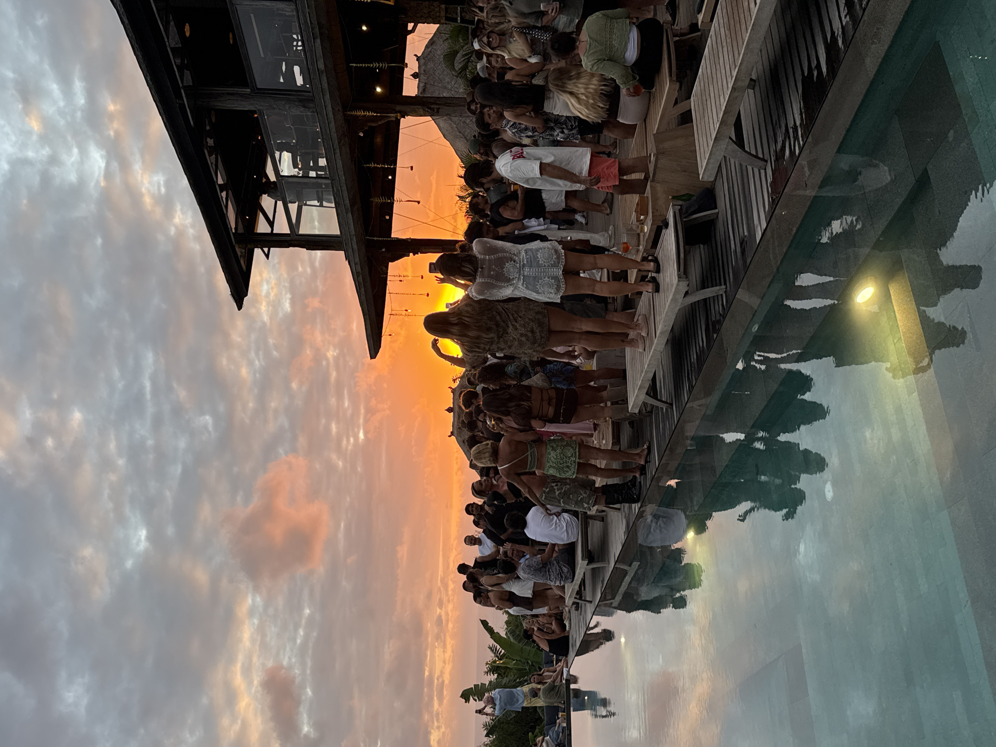 This image depicts a vibrant gathering of people at a waterside venue during sunset. The scene is dominated by a warm, orange glow from the setting sun, which illuminates the sky and casts reflections on the calm water of what appears to be a swimming pool or infinity pool. A multi-level wooden structure, possibly a restaurant or bar, is visible on the left, with its dark timber framing contrasting against the bright sky.
A diverse group of individuals, dressed in casual attire suited for a warm climate, are seen socializing and standing on the wooden deck. Some are looking out towards the sunset, while others appear to be engaged in conversation. The overall mood seems festive and relaxed, with people enjoying the scenic view and each other's company.
Notable details include the rich colors of the sunset, with hues of orange, pink, and gray in the clouds. The water's surface is smooth, reflecting the sky and the silhouettes of the people and structure above. The presence of palm trees in the background hints at a tropical location. The time of day is clearly sunset, given the lighting conditions and the vibrant sky.
While no explicit text is visible, the context provided suggests a location in Kuta Selatan, Indonesia, which is known for its beautiful beaches and sunset views, making this scene characteristic of a popular tourist destination.