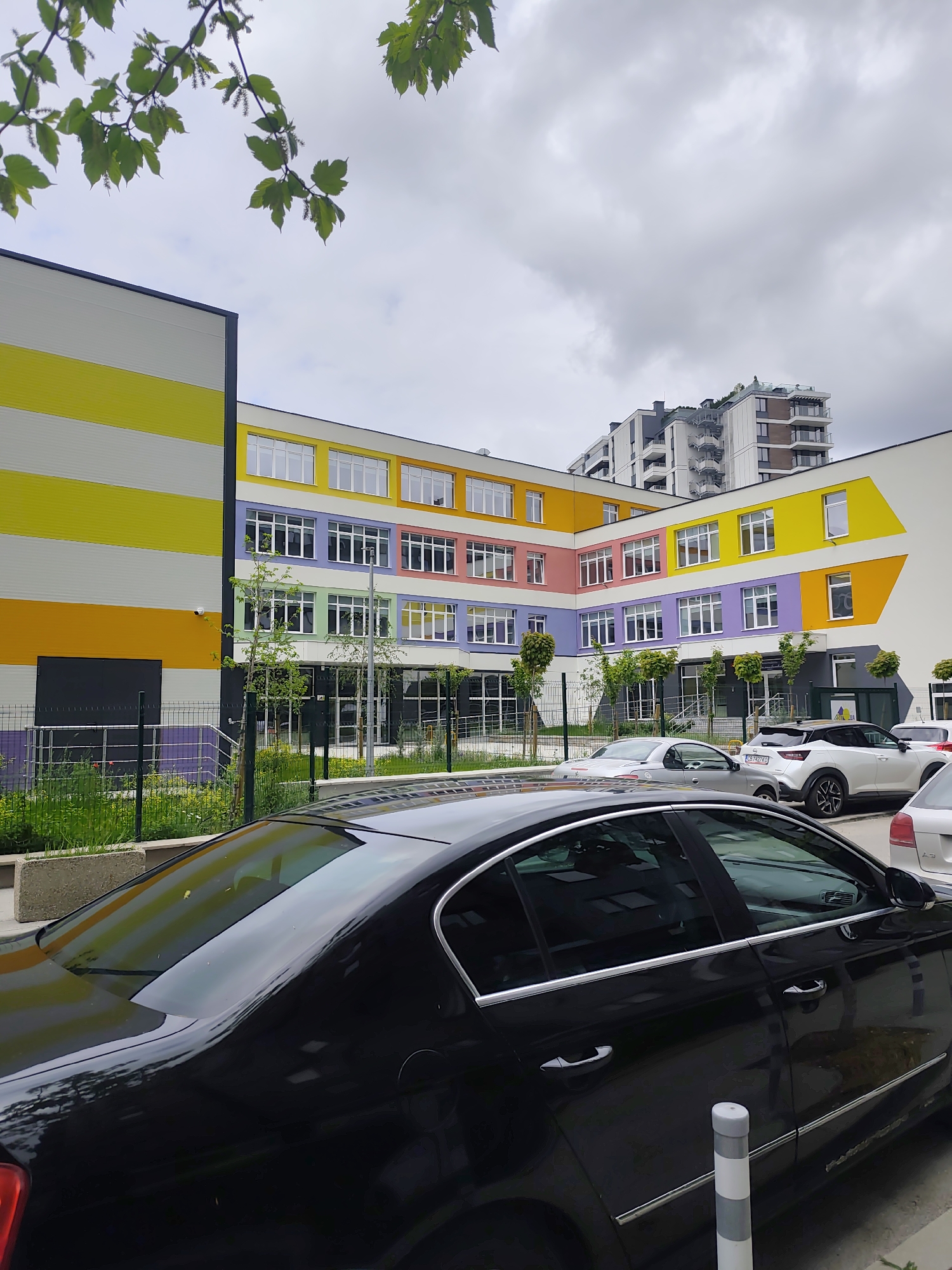 The image shows a modern, brightly colored building with a yellow, white, orange, purple, green, and pink facade. The building has multiple windows and is located next to a parking area with several cars. A black car is parked prominently in the foreground. A taller apartment building stands in the background under a cloudy sky. Trees and vegetation are visible along the fence line and in the foreground. The city and specific location are unknown.
