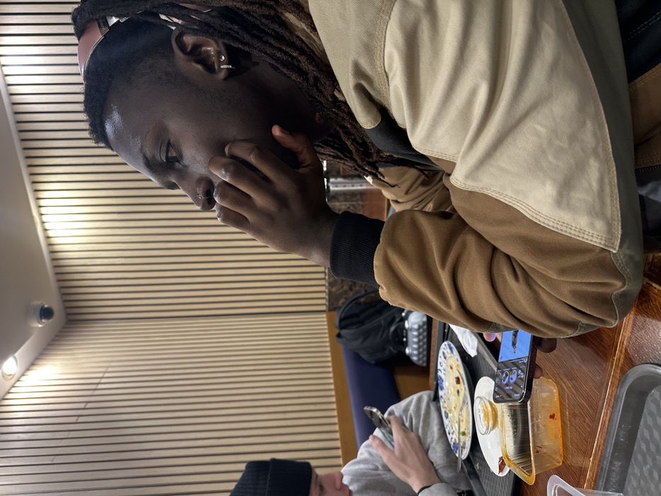 A dark-skinned person with dreadlocks is seated at a table, holding their hand to their face in thought. The person is wearing a two-tone jacket. The table has plates of partially eaten food and a mobile phone propped up on a clear plastic container. Another person wearing a black beanie and grey sweatshirt is seated to the side, looking at their own phone. Overhead are linear ceiling lights, and a security camera is mounted on the wall.