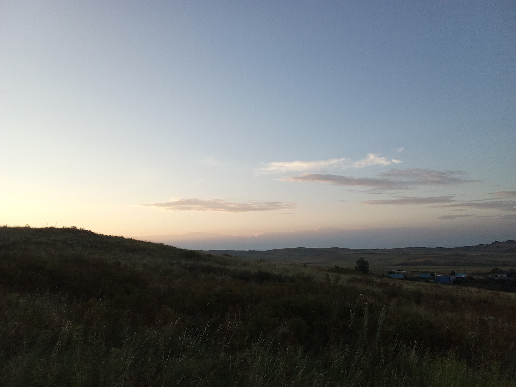 A wide, horizontal shot captures a vast, rolling landscape under a clear, pale blue sky transitioning to soft hues of yellow and pink near the horizon. The foreground is dominated by a dark, undulating grassy hillside, with tall stalks of dry grass catching the light. In the middle ground, a cluster of simple, low-slung buildings with blue roofs is visible amidst the fields, suggesting a small rural settlement. Beyond these structures, the terrain continues to roll into the distance, with subtle undulations and a single, dark tree standing to the left of the buildings. The overall impression is one of a tranquil, open rural environment in Samsonovka, Kazakhstan, likely at dawn or dusk due to the soft lighting.