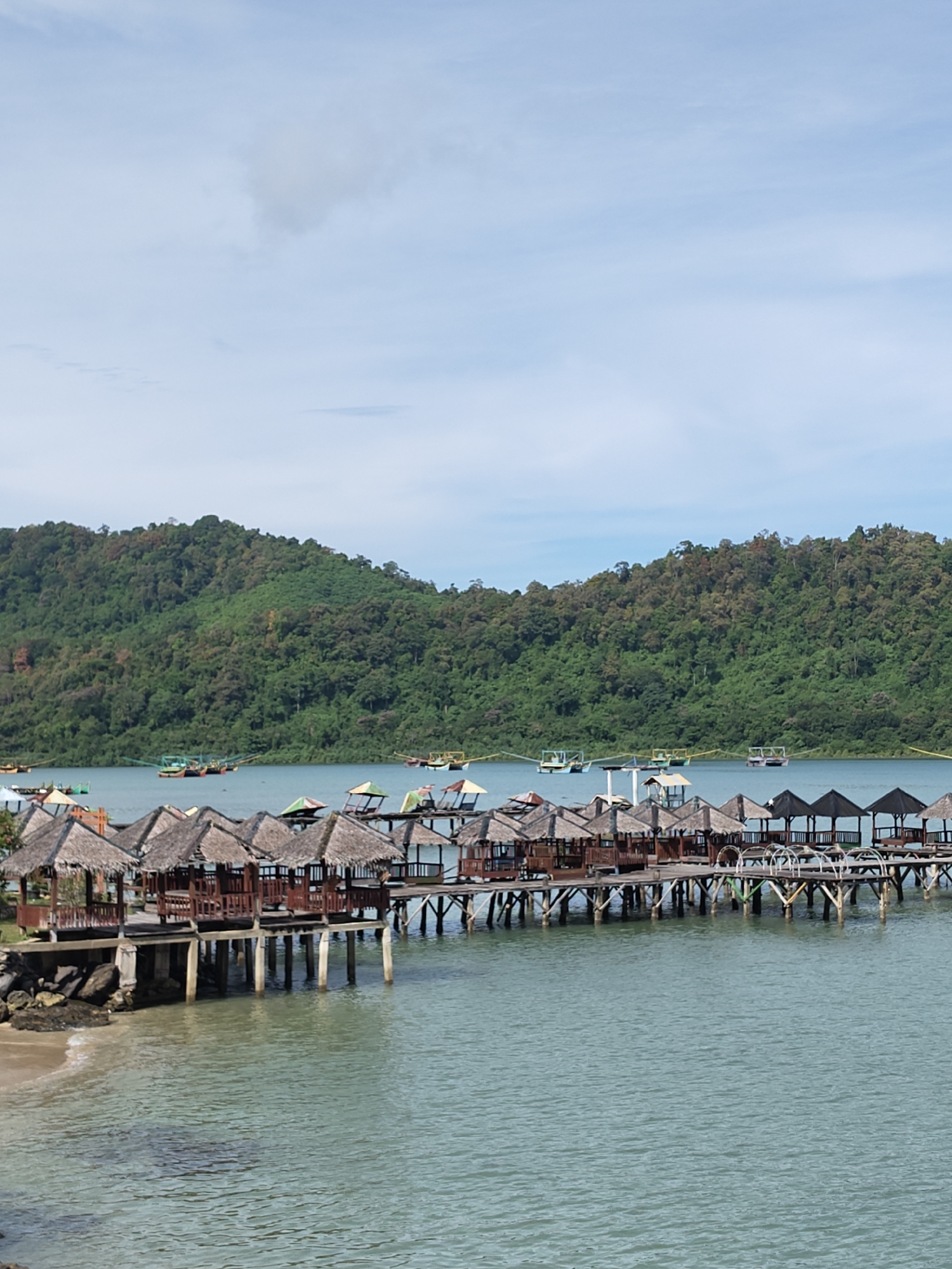 The image depicts a waterfront scene in Layeun, Indonesia. A long pier with huts extends into the water. The huts have thatched roofs and are supported by wooden pillars. In the background, there's a forested hill and several boats on the water. The sky is mostly blue with a few clouds. The water is a light blue-green color.