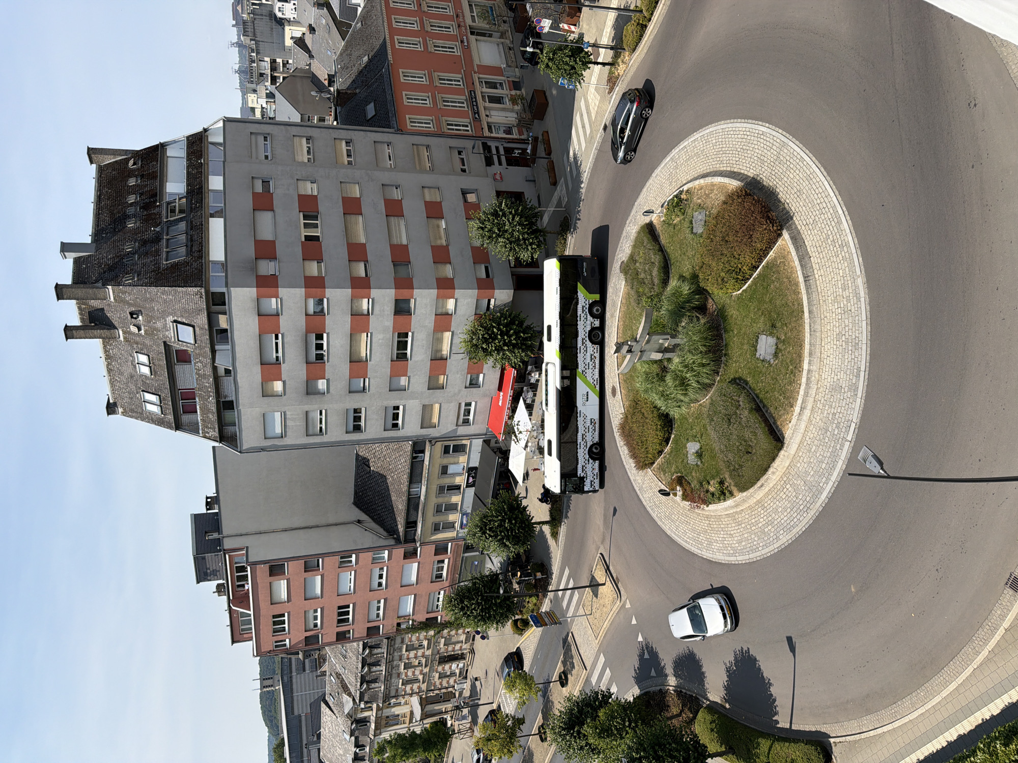 The aerial view captures a sunny daytime scene at a roundabout in Esch-sur-Alzette, Luxembourg. A modern, multi-story building with a grey facade and red rectangular window accents dominates the background. To its left, a taller, reddish-pink apartment building features numerous white-framed windows. The roundabout itself is circular with a landscaped central island containing low-lying shrubs and grasses.
Several vehicles are present: a white car is entering the roundabout from the lower left, and a dark-colored hatchback is exiting on the right. A long, green and white bus with a patterned design is in motion, traversing the roundabout. Streetlights are visible along the road, and trees line the surrounding sidewalks and the roundabout's edge. The architecture suggests a mixed urban environment with residential and commercial structures. The sky is clear and blue, indicating fair weather.