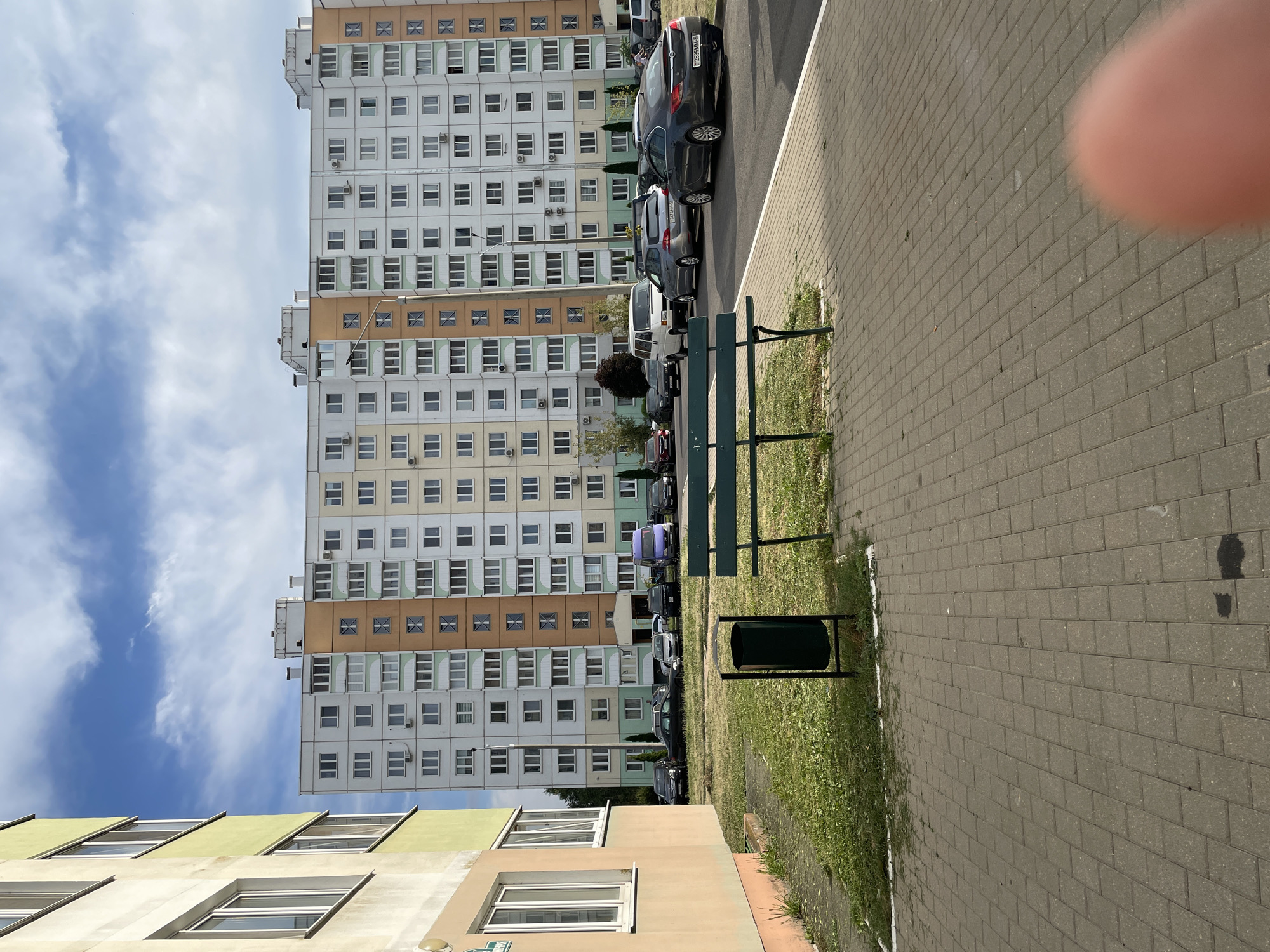 An exterior daytime, eye-level shot shows a residential area in Salihorsk, Belarus. The foreground features a paved walkway and a green metal bench, with a dark green trash can located to the left of the bench on a patch of grass. Behind the bench, a large, multi-story apartment building dominates the background. The building has a colorful facade with sections of white, light green, and orange. Numerous windows are visible on each level. A line of parked cars, including a white van and several sedans, are situated in front of the apartment building. The sky above is blue with scattered white clouds. The overall scene depicts a typical urban residential environment.