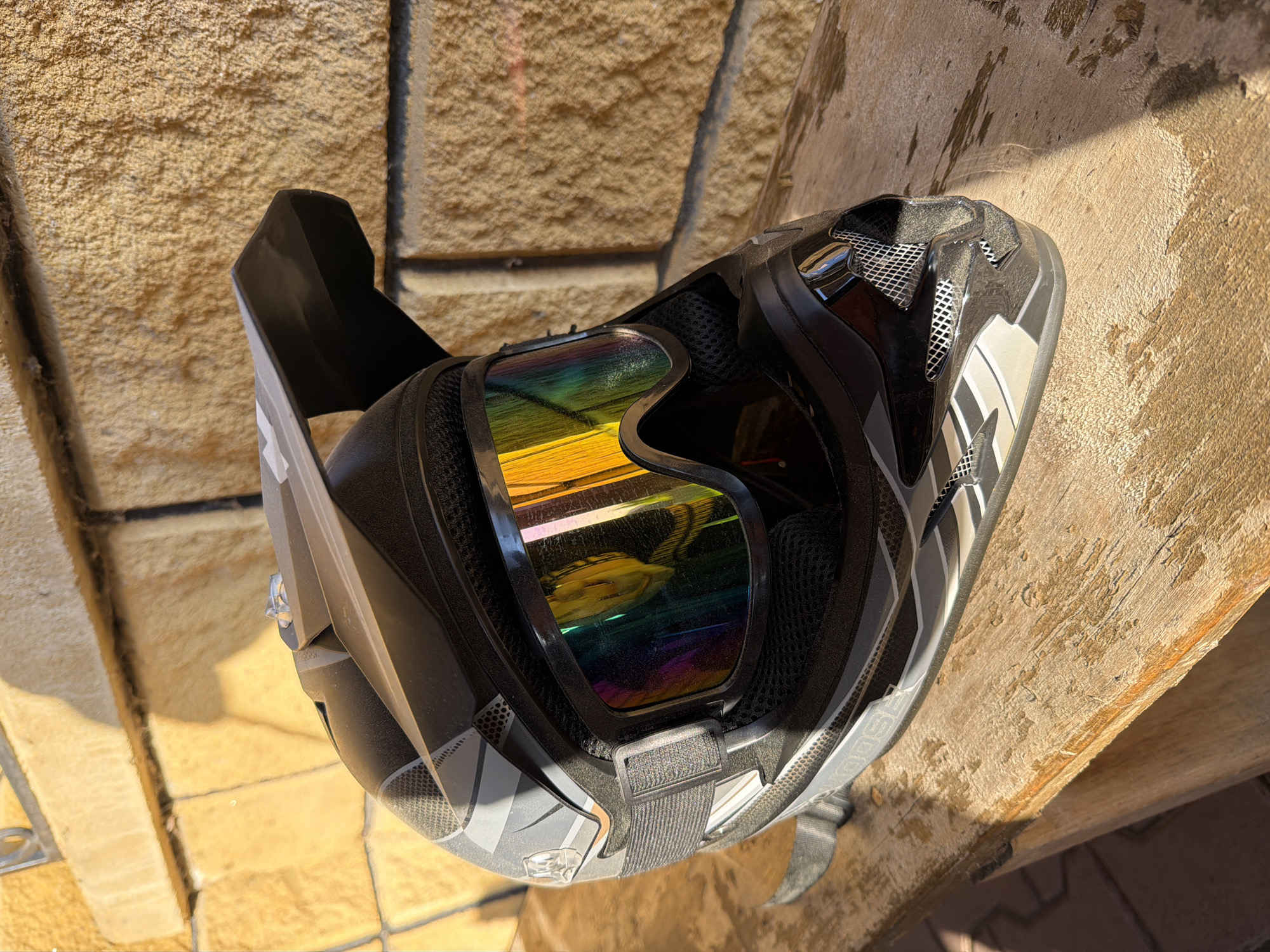 A gray and black dirt bike helmet with rainbow-colored goggles inside sits on a rough, light-brown wooden surface. The background consists of a wall made of light-brown, textured stone bricks. The scene is lit by bright, direct sunlight. There is no evidence indicating this is Bolsheustyikinskoye, Russia.