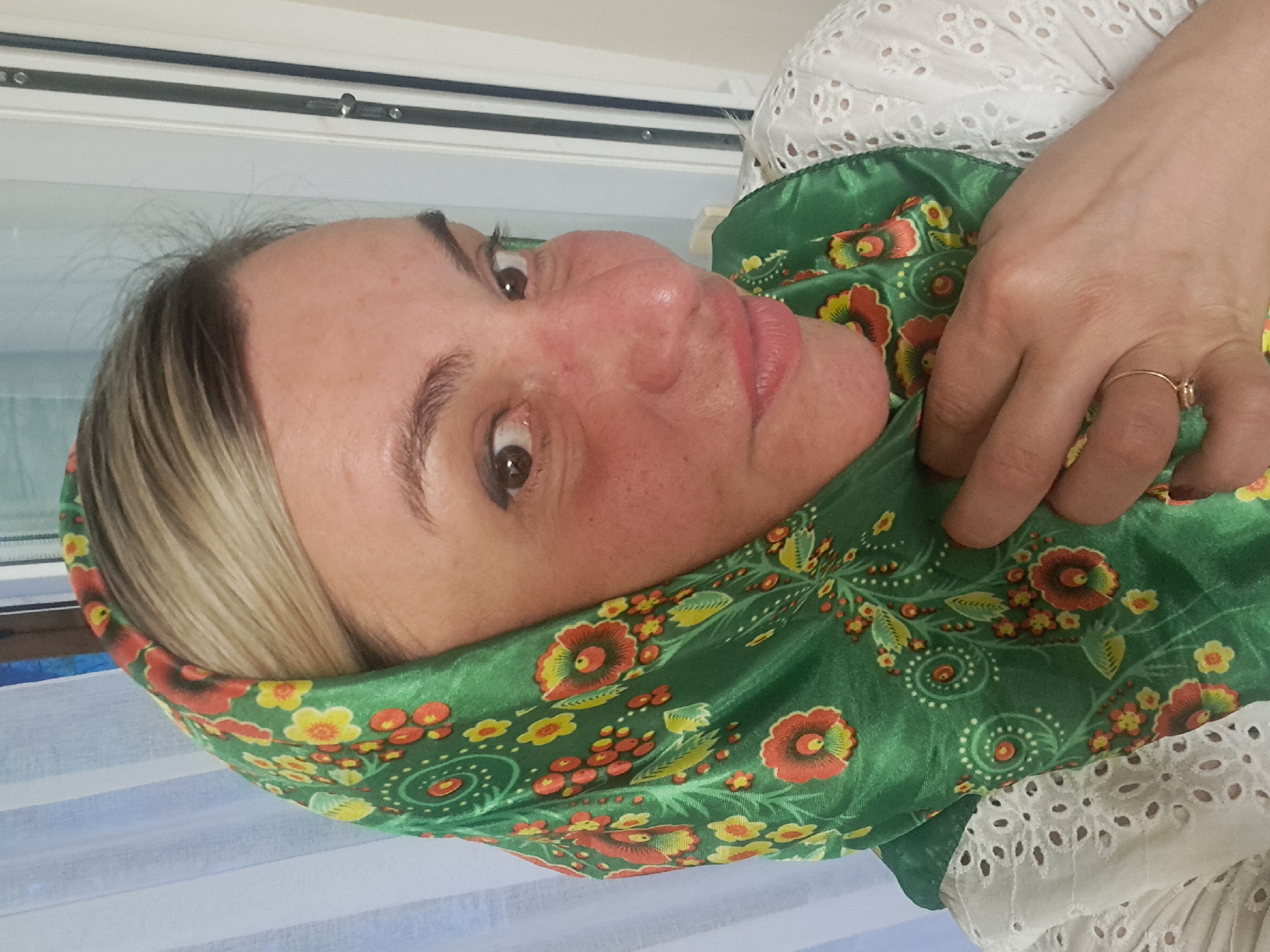 A woman with fair skin, blonde hair, and brown eyes is wearing a green floral scarf and a white eyelet top. She is indoors near a window. It is not possible to determine the location in the city of Elektrostal, Russia, from the image alone.