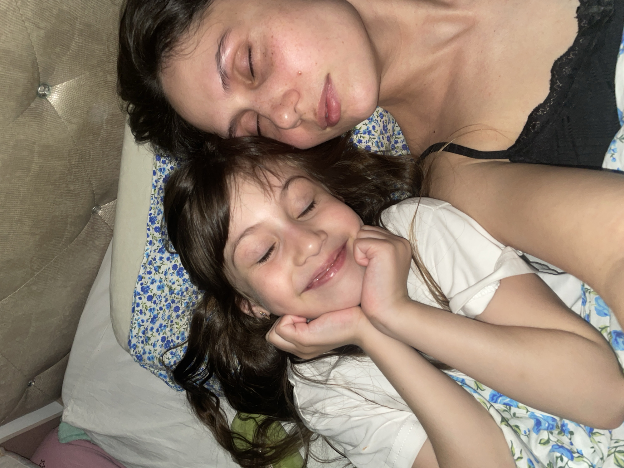 A close-up, high-angle shot captures two females, likely a mother and daughter, resting peacefully side-by-side in what appears to be a bed. The scene evokes a sense of intimacy and tranquility.
The older female, positioned in the upper part of the frame, has dark hair cascading around her head. Her eyes are gently closed, and her facial expression is serene. She has fair skin, with visible freckles across her nose and cheeks. She is wearing a dark top, possibly a black bra or camisole, with delicate lace detailing visible on the strap and upper edge. Her body is oriented horizontally, with her head resting on a pillow.
Below and slightly in front of the older female is a younger girl. She has long, dark brown hair spread out around her. Her eyes are also closed, but a broad, gentle smile plays on her lips, suggesting a state of contentment, happiness, or a pleasant dream. Her chin is resting on her clasped hands, which are positioned just below her face. She is wearing a simple white short-sleeved t-shirt.
The two individuals are lying on patterned bedding, primarily featuring a white fabric with a repeating blue floral motif and green leaves. A white pillow is also visible. In the background, to the left of the older female's head, there's a padded headboard in a light brown or taupe color, adorned with decorative metallic studs or buttons. The soft, somewhat dim lighting indicates it could be evening, night, or early morning. The overall atmosphere is one of peaceful slumber and affection within a home setting, possibly in Izhevsk, Russia. There is no visible text in the image.