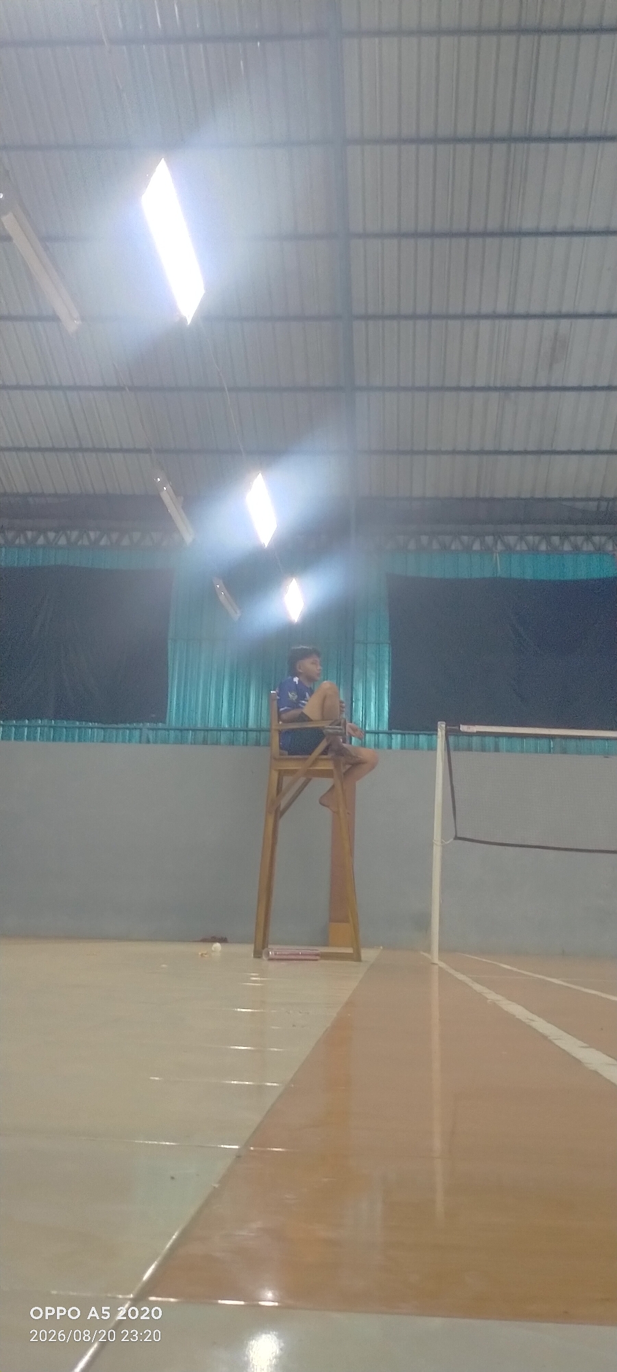 A young male, appearing to be a teenager, is seated on a high wooden chair on a badminton court. He is wearing a blue and white athletic shirt and brown shorts. The chair is positioned near the center of the court, next to the net. The court has distinct light and dark brown markings. Above, the corrugated metal ceiling of the indoor facility is visible, with bright fluorescent lights illuminating the space. The background consists of a gray wall and a dark curtain. The timestamp in the bottom left corner indicates the image was taken on August 20, 2026, at 23:20. The location is identified as Dongos, Indonesia.