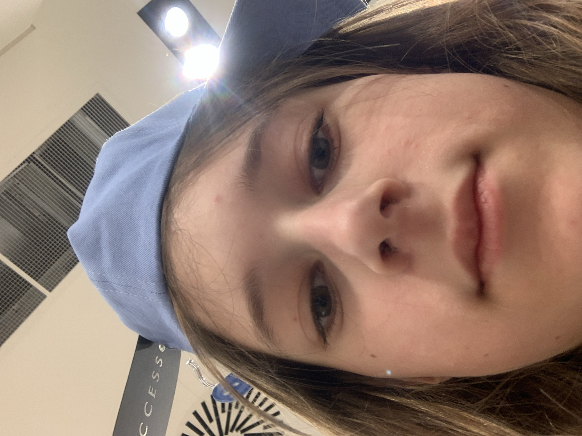 This close-up, upward-angled selfie features a young person, likely female, captured indoors in what appears to be a commercial or public building in Belgrade, Serbia. The subject has light brown hair framing her face and is wearing a light blue cap or a light blue hoodie. Her eyes, appearing to be a dark blue or greyish-green, are mostly directed towards the camera, while her expression is neutral with a slight hint of a smile on her lips. A small, metallic earring is visible near her right ear.
The background reveals a light-colored ceiling with prominent, bright artificial overhead lights causing lens flare, particularly near the top of the subject's head. Part of a ventilation grille or an air conditioning unit is visible on the ceiling. Below the subject's right ear, a black rectangular object, possibly a retail tag or a sign, is partially visible with white text that reads "CCESS E". The lighting suggests an indoor setting, and the nature of the background elements points to a public space such as a mall or a store. The image's orientation indicates it was taken from a low angle, with the camera possibly tilted.