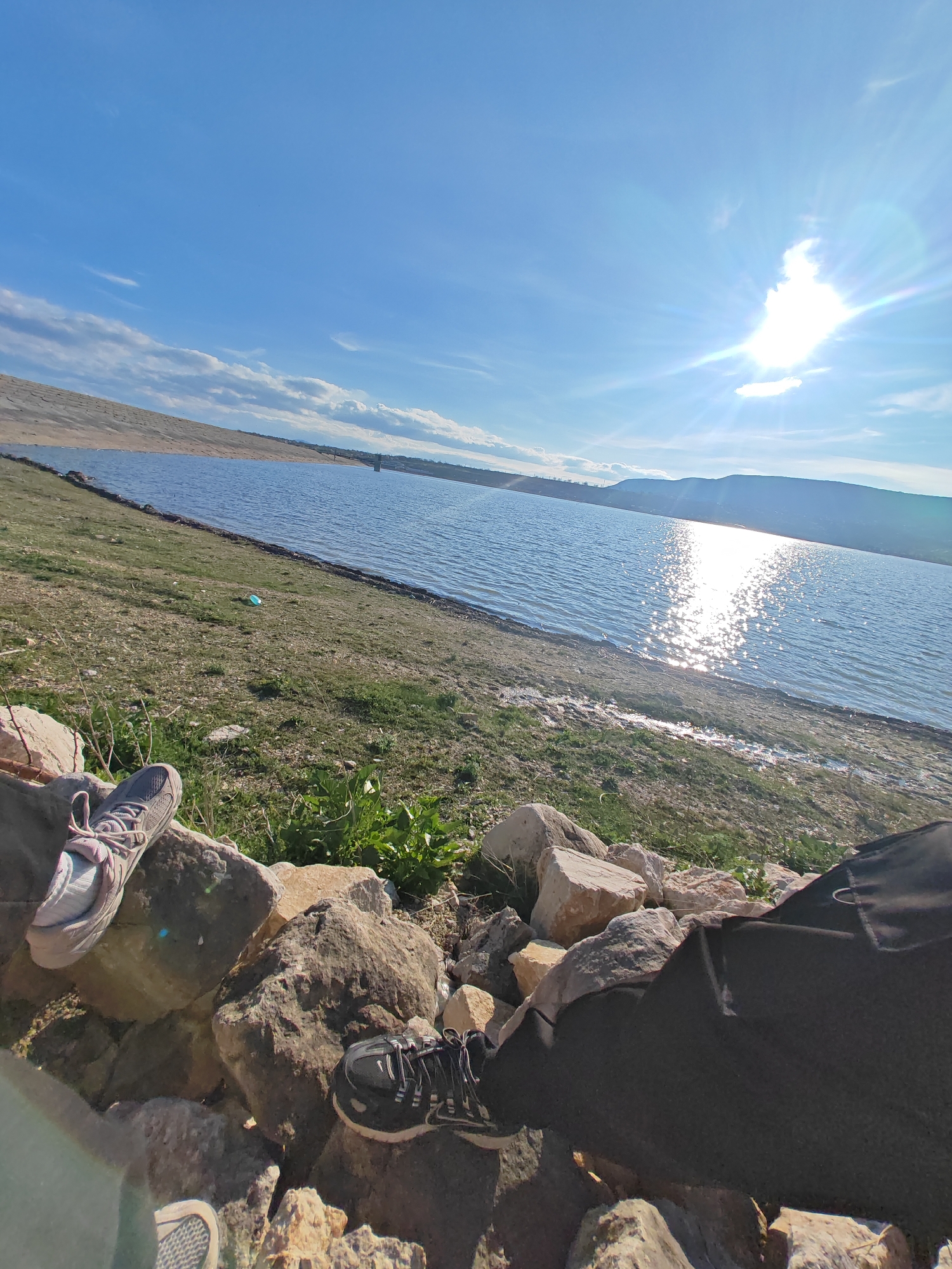 The image shows a seated person's perspective on a sunny day by a lake or reservoir. The person is sitting on a rocky area at the water's edge, with their legs and shoes visible in the foreground. There are two pairs of athletic shoes visible. The landscape includes grass and vegetation along the shore, with a body of water stretching into the distance. The sun is bright, reflecting on the water's surface. Hills or mountains are visible on the opposite shore. The sky is blue with scattered clouds. The city of Shumen, Bulgaria is not visually identifiable in the image.