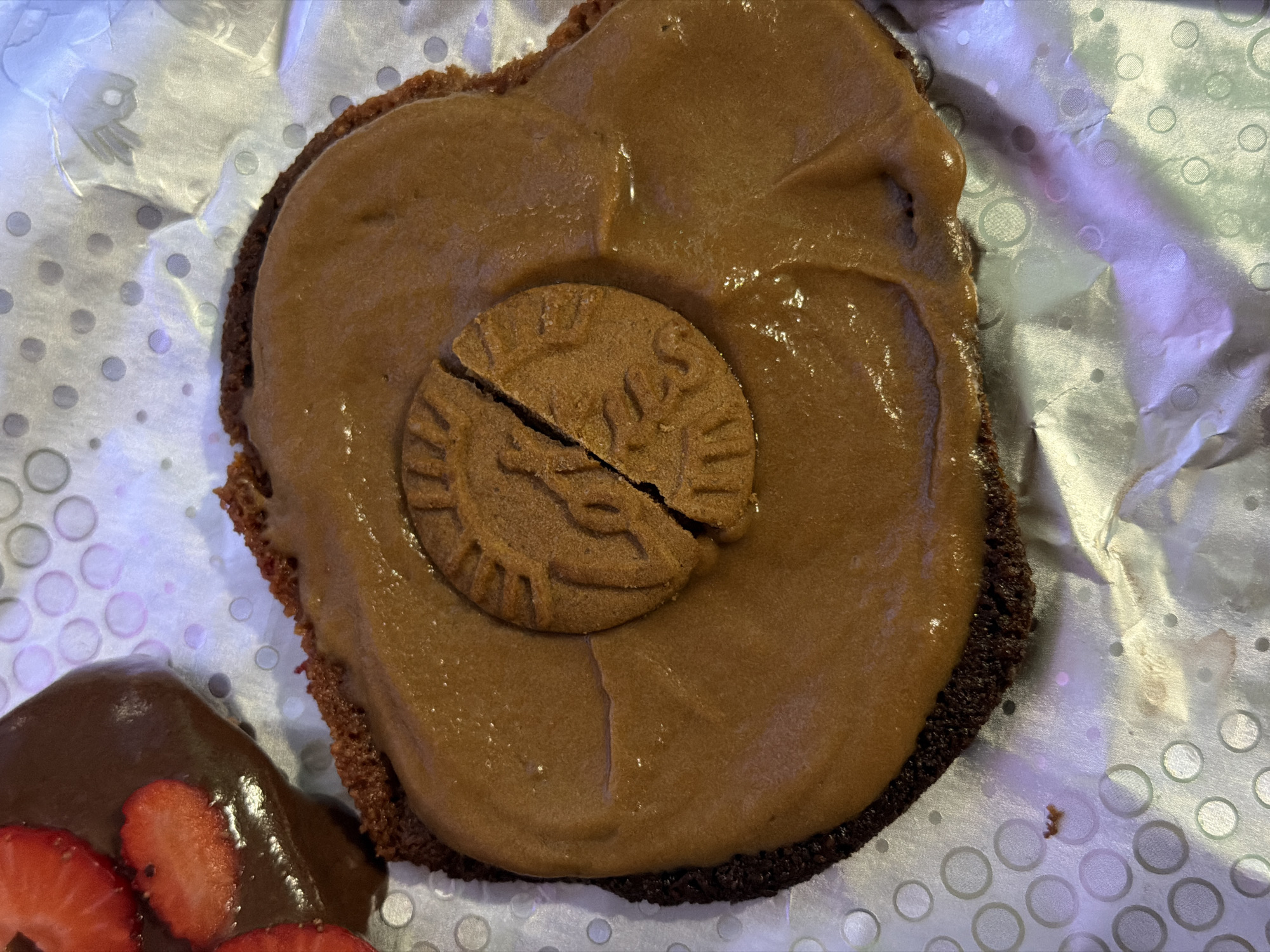 A close-up shot depicts a dessert item resting on a patterned silver surface. The dessert consists of a brown base layer, topped with a smooth, light brown frosting. A broken, circular cookie is positioned on the frosting. In the lower-left corner, a portion of another dessert is visible, featuring chocolate sauce and sliced strawberries. The silver surface has a pattern of small circles. The location is not identifiable from the image.