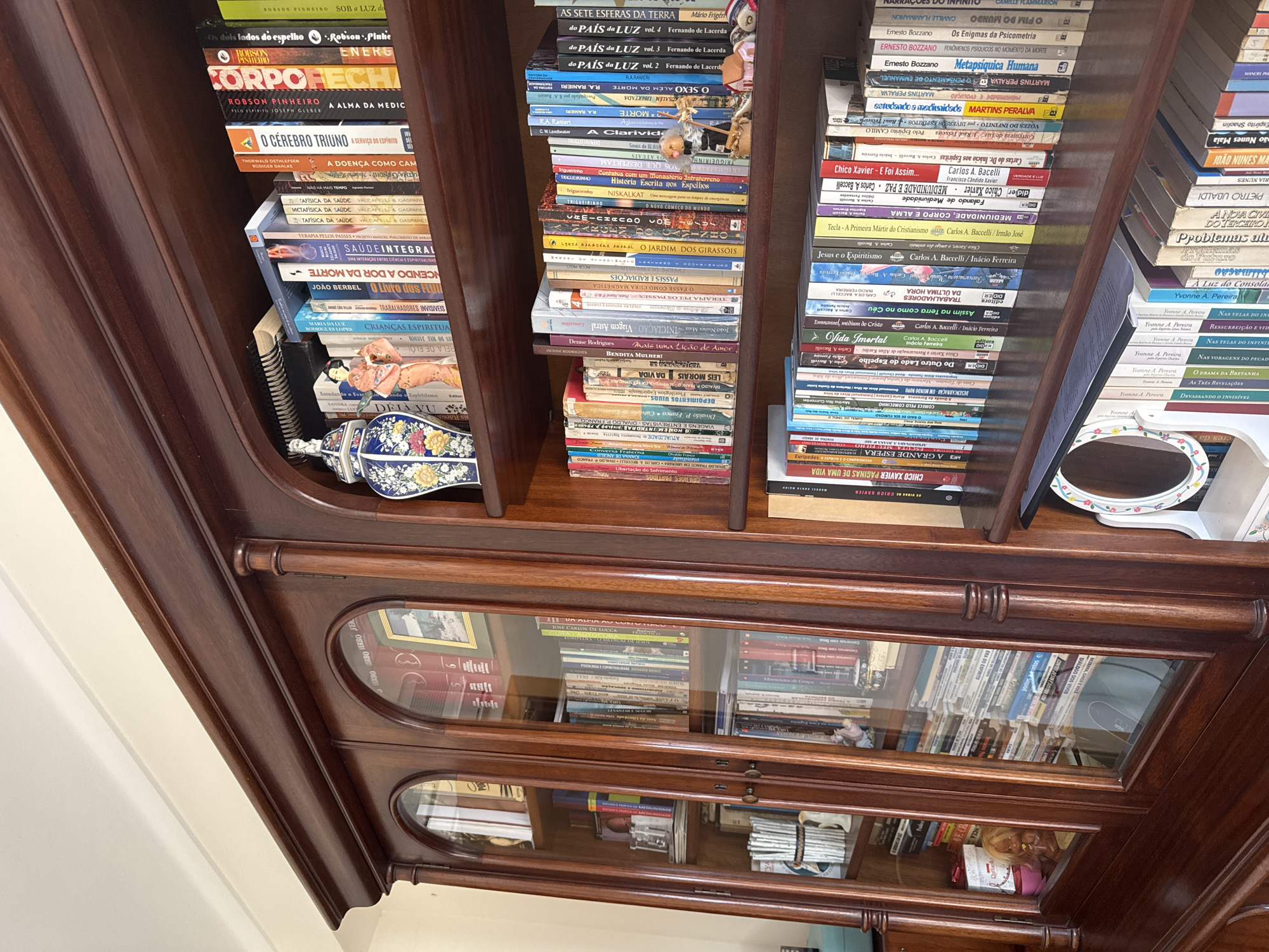 The image displays an interior scene in Mirandela, Portugal, focused on a large, dark reddish-brown wooden display cabinet and bookshelf. The furniture features a classical design with decorative molding, fluted pilasters, and two primary sections. The left section functions as a cabinet, featuring four arch-topped glass doors that enclose multiple shelves. Visible items behind the glass include numerous books, stacks of white tubular objects, a small white figurine, and a blonde doll in a pink top positioned next to a pink cup on a lower shelf.
The right section comprises open bookshelves with four visible shelves, each densely filled with books, primarily in Portuguese, along with various decorative objects. The top open shelf features a blue and white floral-patterned ceramic vase on the left, followed by an orange-clad Asian figurine, and numerous books with titles such as "O CÉREBRO TRIVUNO," "CORPO É FÉ," and "METAFÍSICA DA SAÚDE." The second open shelf below contains additional books and a small figurine depicting an elderly person in brown attire seated next to miniature house structures. The third open shelf from the top displays more books, with titles including "CHICO XAVIER PÁGINAS DE UMA VIDA," "VIDA IMORTAL," and "MEDIUNIDADE, CORPO E ALMA," attributed to authors like Carlos A. Baccelli and Chico Xavier. The partially visible bottom open shelf on the right contains more books and a white, oval-shaped vanity mirror with a floral pattern. The background consists of a plain, light-colored wall and ceiling. No individuals are present in the scene.