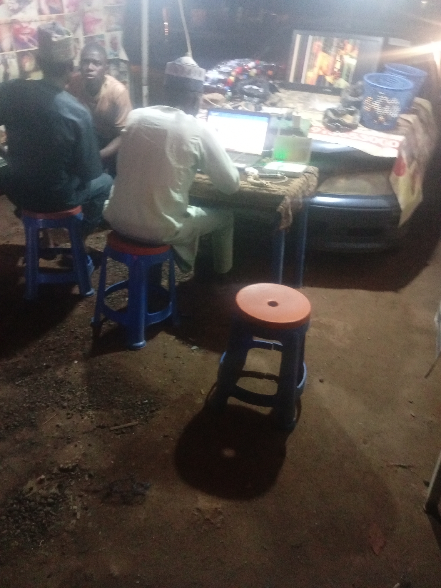 In the city of Tundun Wada West, Nigeria, this nighttime scene depicts an outdoor setting with a table laden with items, including a laptop and a television monitor. Three individuals are present, with two seated on blue stools and one standing. The lighting appears to be artificial, possibly from a nearby lamp. The ground is uneven and appears to be dirt with scattered small stones. In the background, a vehicle is partially visible. The general ambiance suggests a casual, communal gathering or a small business operation.